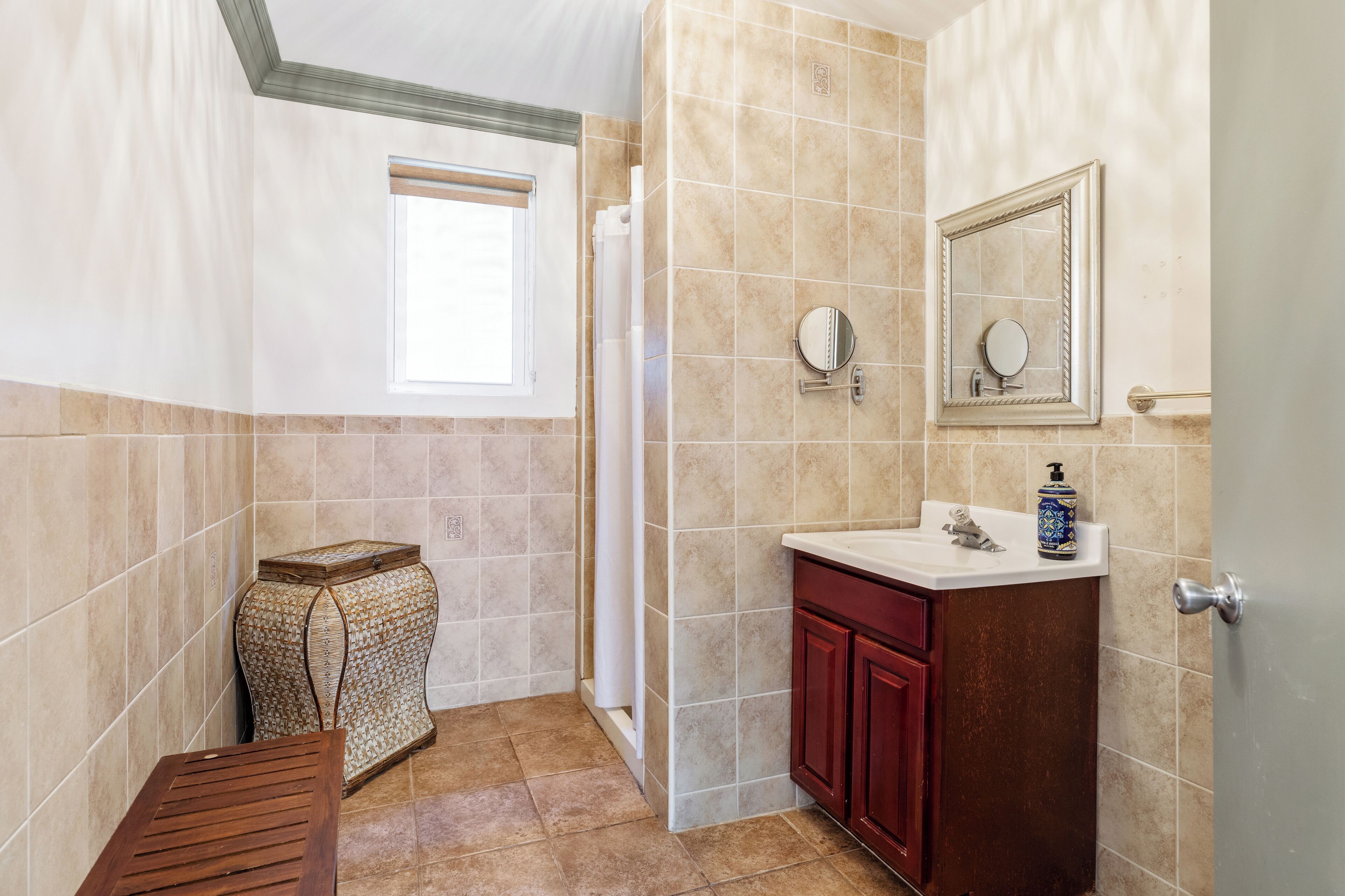 A bathroom with beige tiles, a wooden bench, a vanity with a mirror, and a window.