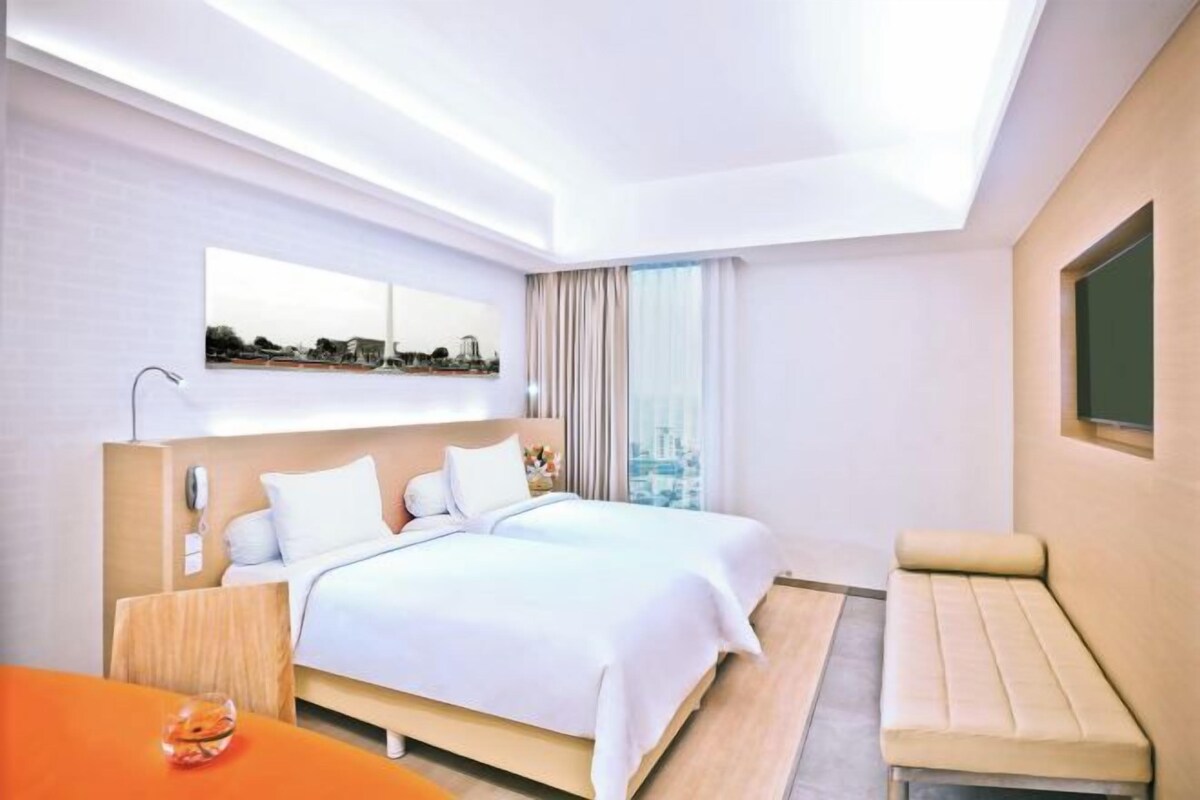 Habitación de hotel con dos camas, zona de estar, vista a la ciudad y un ventanal grande.
