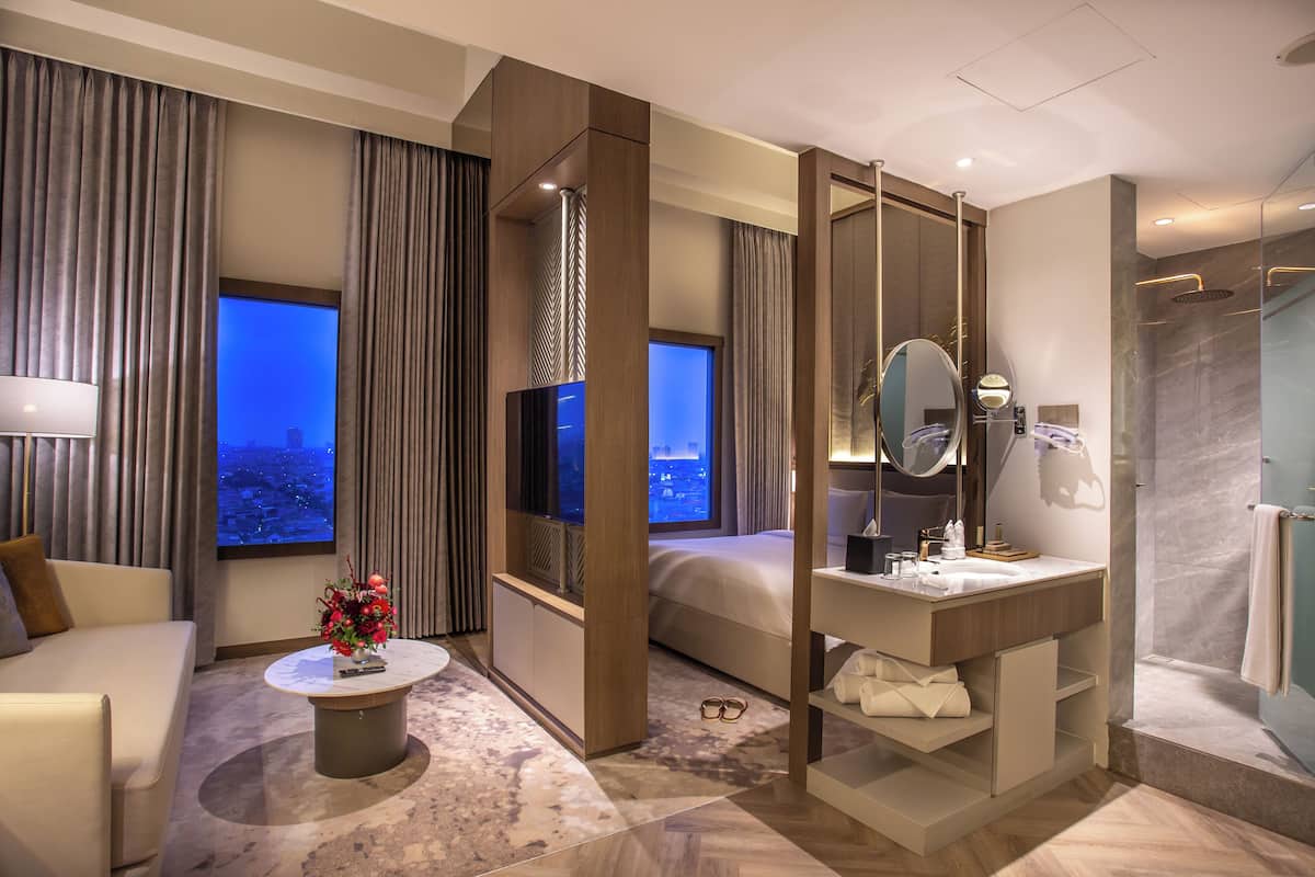 A hotel room with a bed, a sitting area with a coffee table, a bathroom with a mirror and sink, and a view of the city at night.