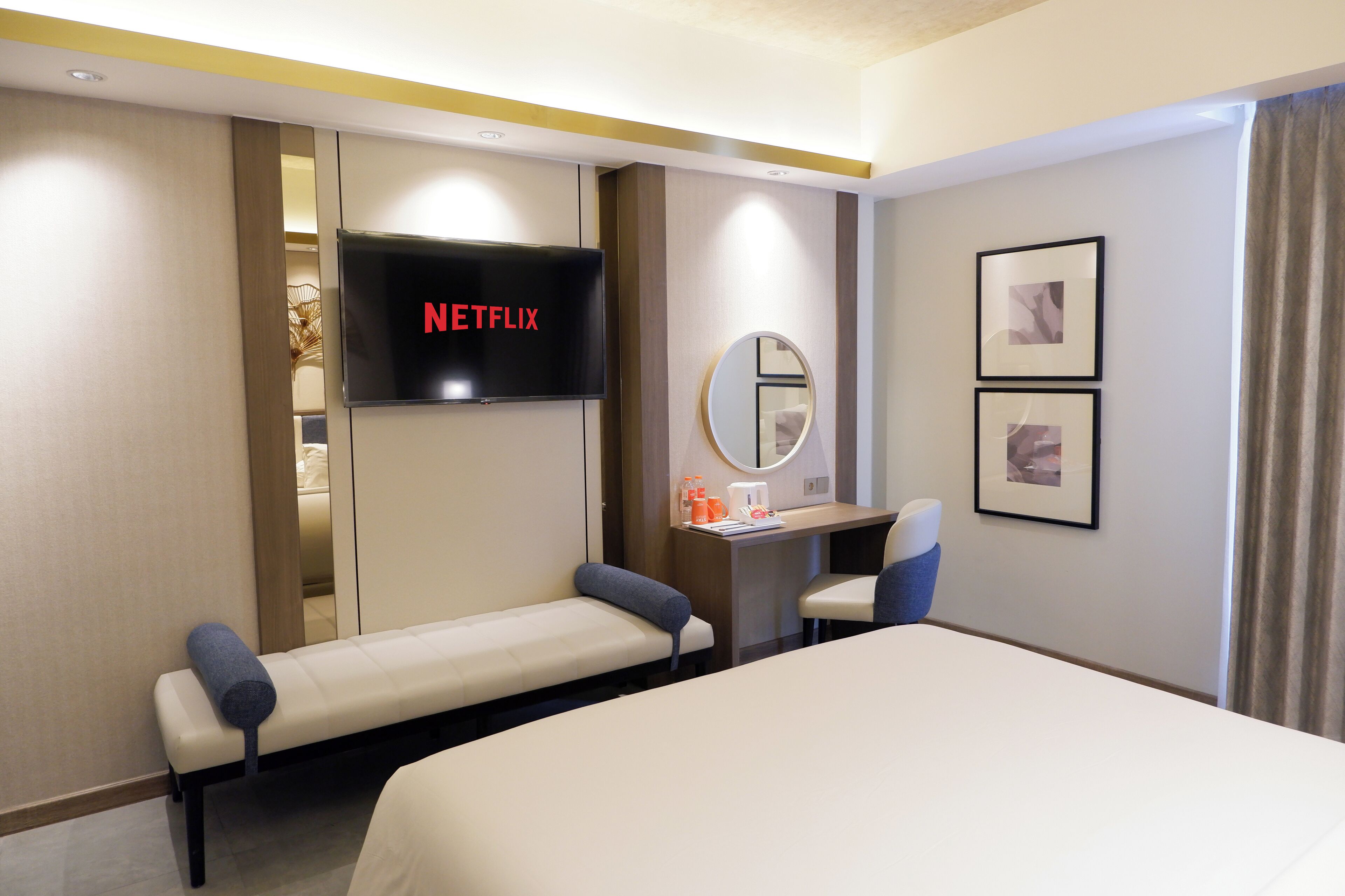 A hotel room with a bed, a desk, a chair, a TV displaying 'NETFLIX', a mirror, and framed pictures on the wall.