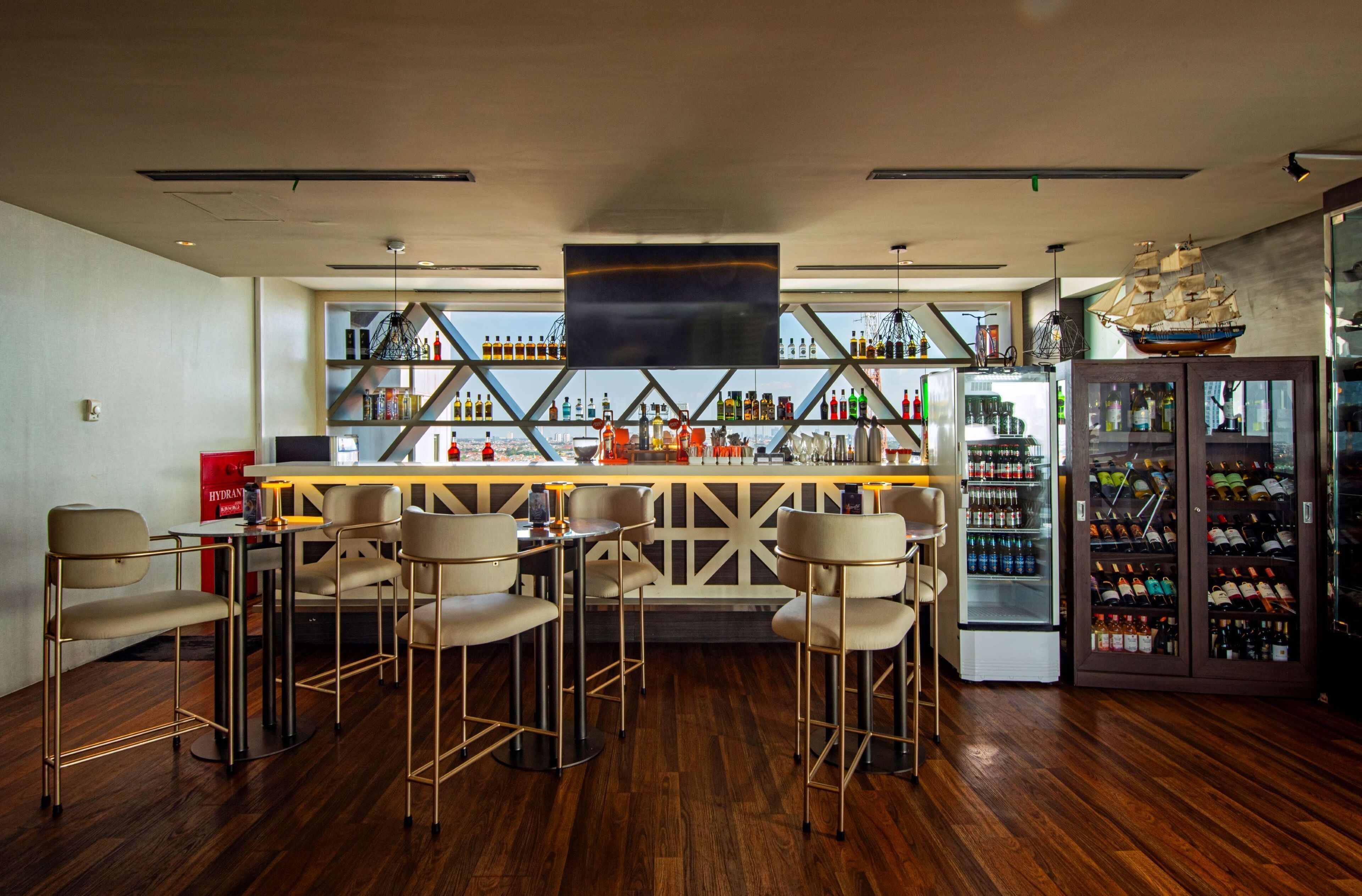 A bar area with wooden flooring, bar stools, a large TV screen, and a wine refrigerator.