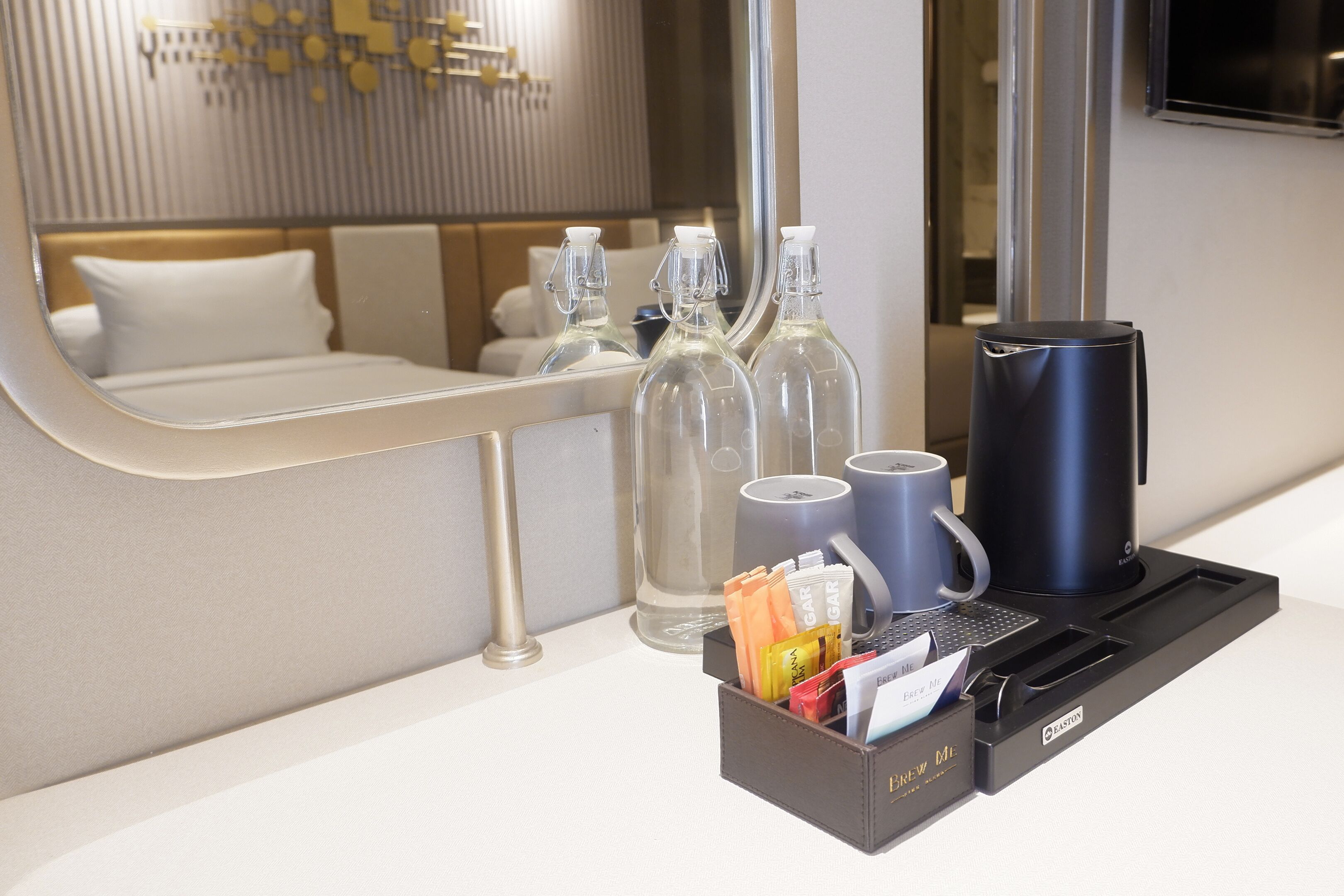 A hotel room with a bed, a bedside table with a coffee maker, a glass bottle, and a black tumbler.