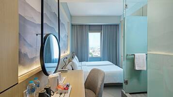 Ett hotellrum med en säng, ett skrivbord med en stol, en spegel, ett fönster med gardiner och en duschkabin med glasväggar.