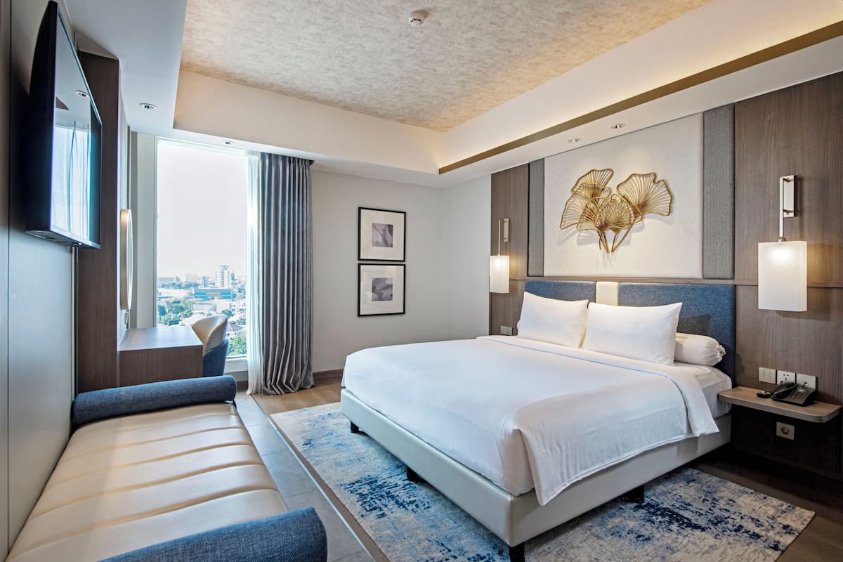 A modern hotel room with a large bed, a seating area, a city view, and a decorative wall piece.