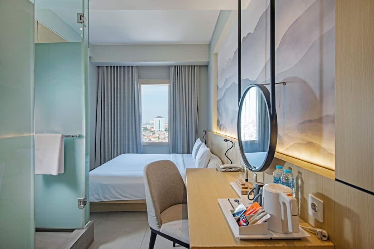A hotel room with a bed, a desk with a mirror, a chair, a window with curtains, and a view of a cityscape.