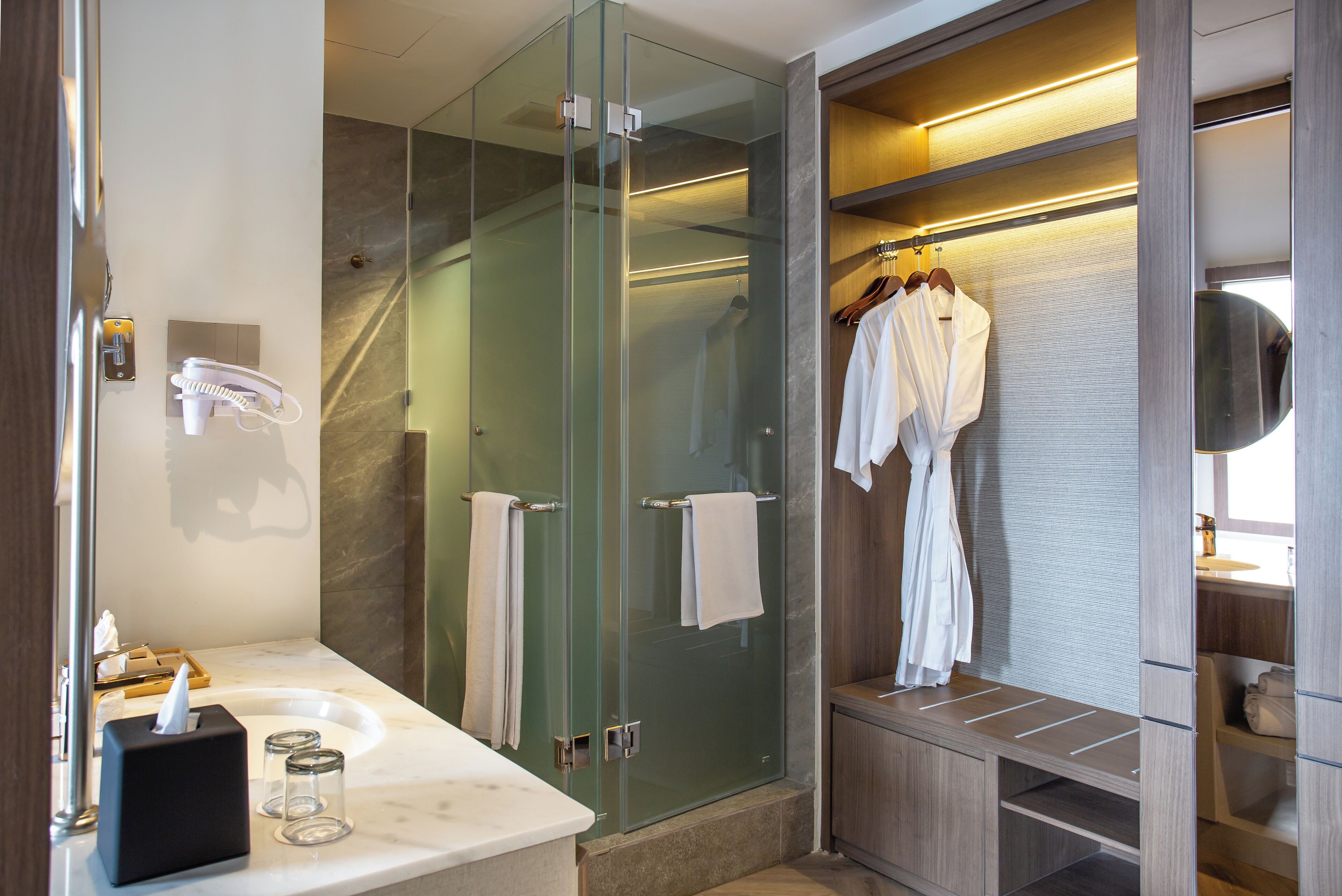 A modern bathroom with a glass-enclosed shower, a vanity with a marble countertop, and a walk-in closet with hanging robes.
