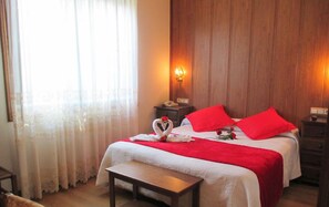 Quarto de hotel com cama, mesa de cabeceira, criado-mudo com abajur e janela com cortinas.