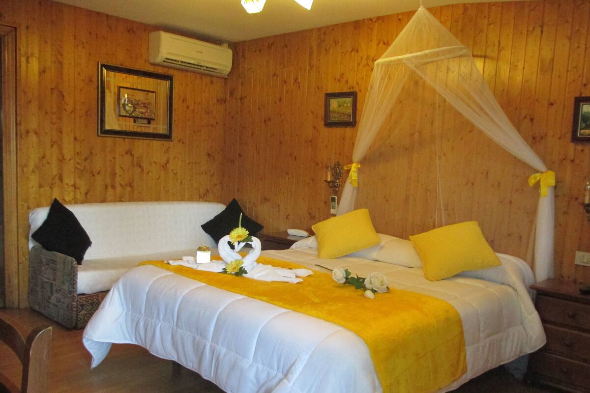 Ein Zimmer mit Holzwandverkleidung, einem Bett mit weißer und gelber Bettwäsche, einem Himmelbett und einem Nachttisch mit Lampe.