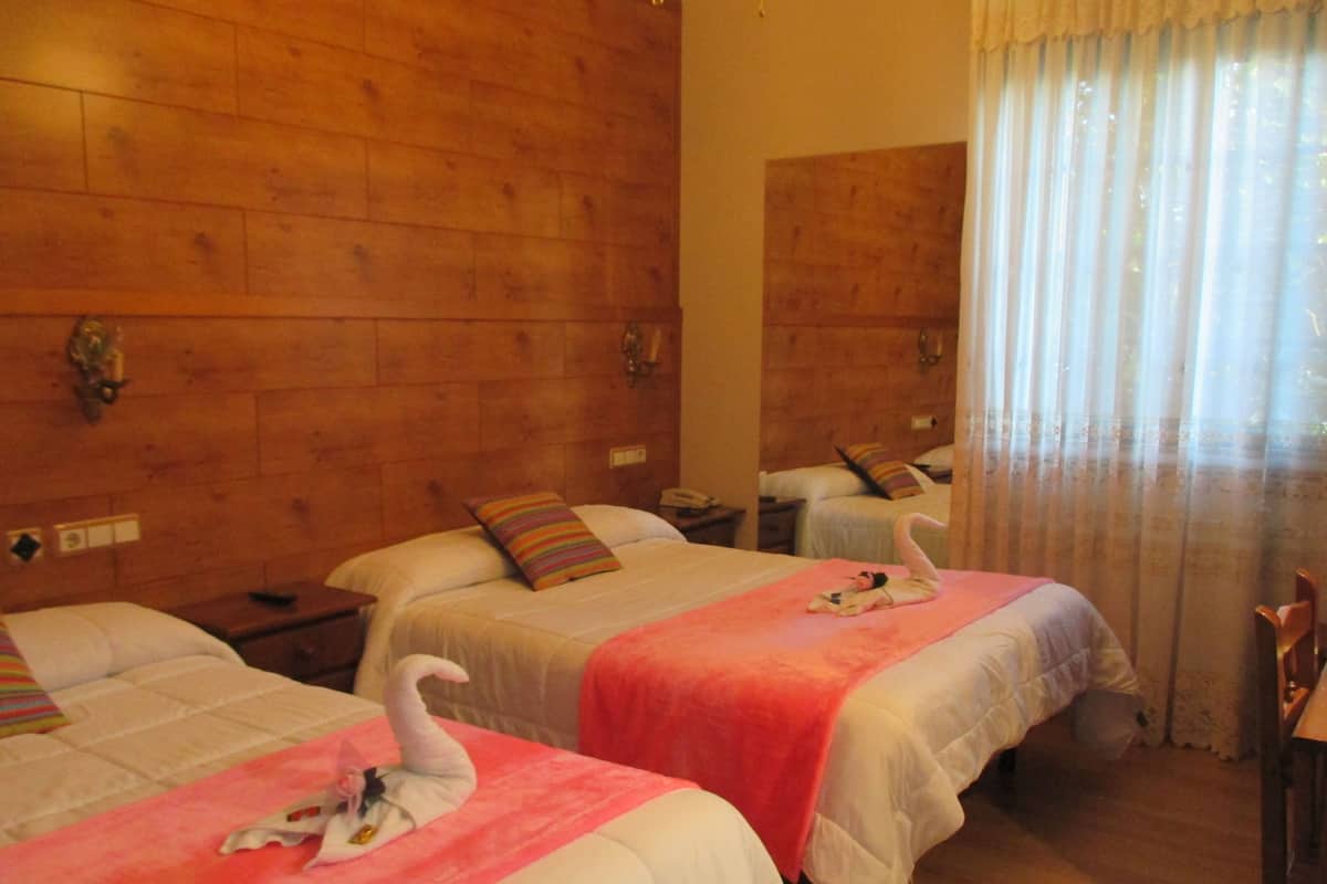 Ein Hotelzimmer mit drei Betten, Holzwandverkleidung, einem Spiegel und einem Fenster mit durchscheinenden Vorhängen.