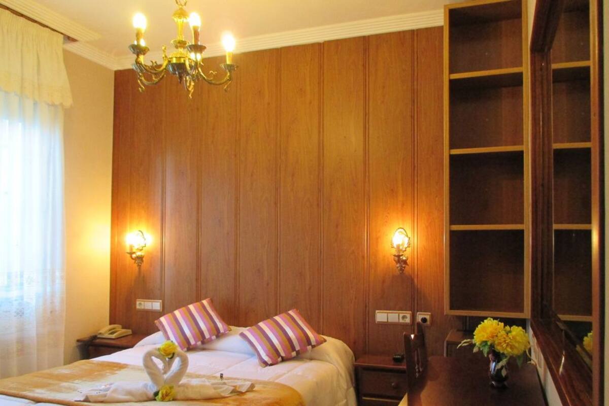 Ein Hotelzimmer mit einer holzverkleideten Wand, einer Kristallleuchter, einem Bett mit gestreiften Kissen, einem Nachttisch mit Lampe und einer Vase mit Blumen.