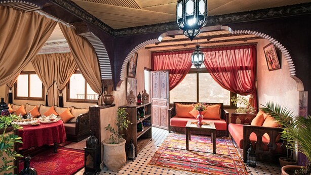 A traditional Moroccan interior with ornate arches, a patterned rug, and a variety of furnishings including sofas and a table.
