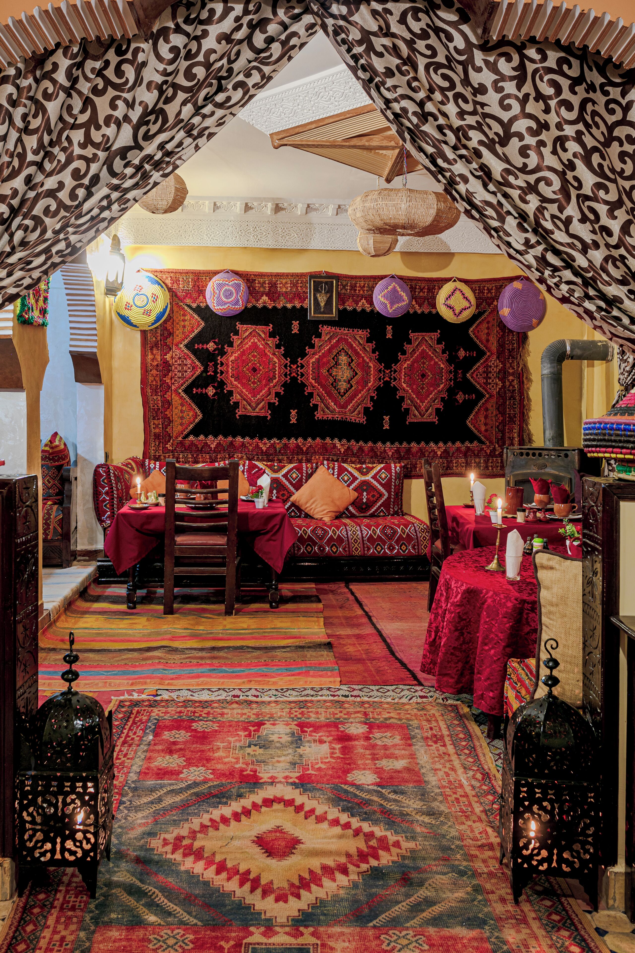 A traditional room with a patterned carpet, a bed with a red cover, a dining table with a candle, and a hanging lantern.
