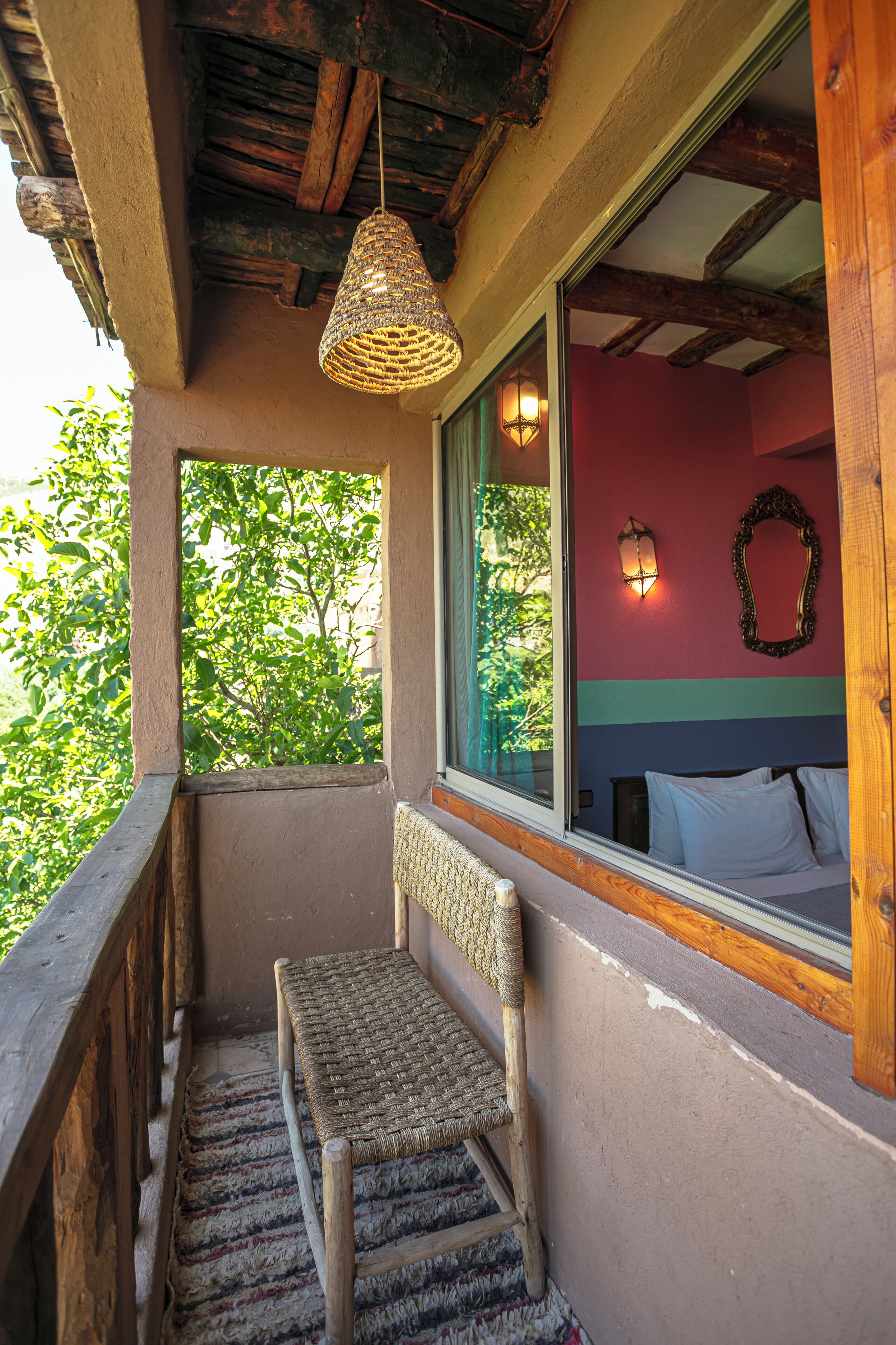 A balcony with a wicker chair, a wooden railing, a window with a view of greenery, and a wall with a mirror and wall-mounted lights.