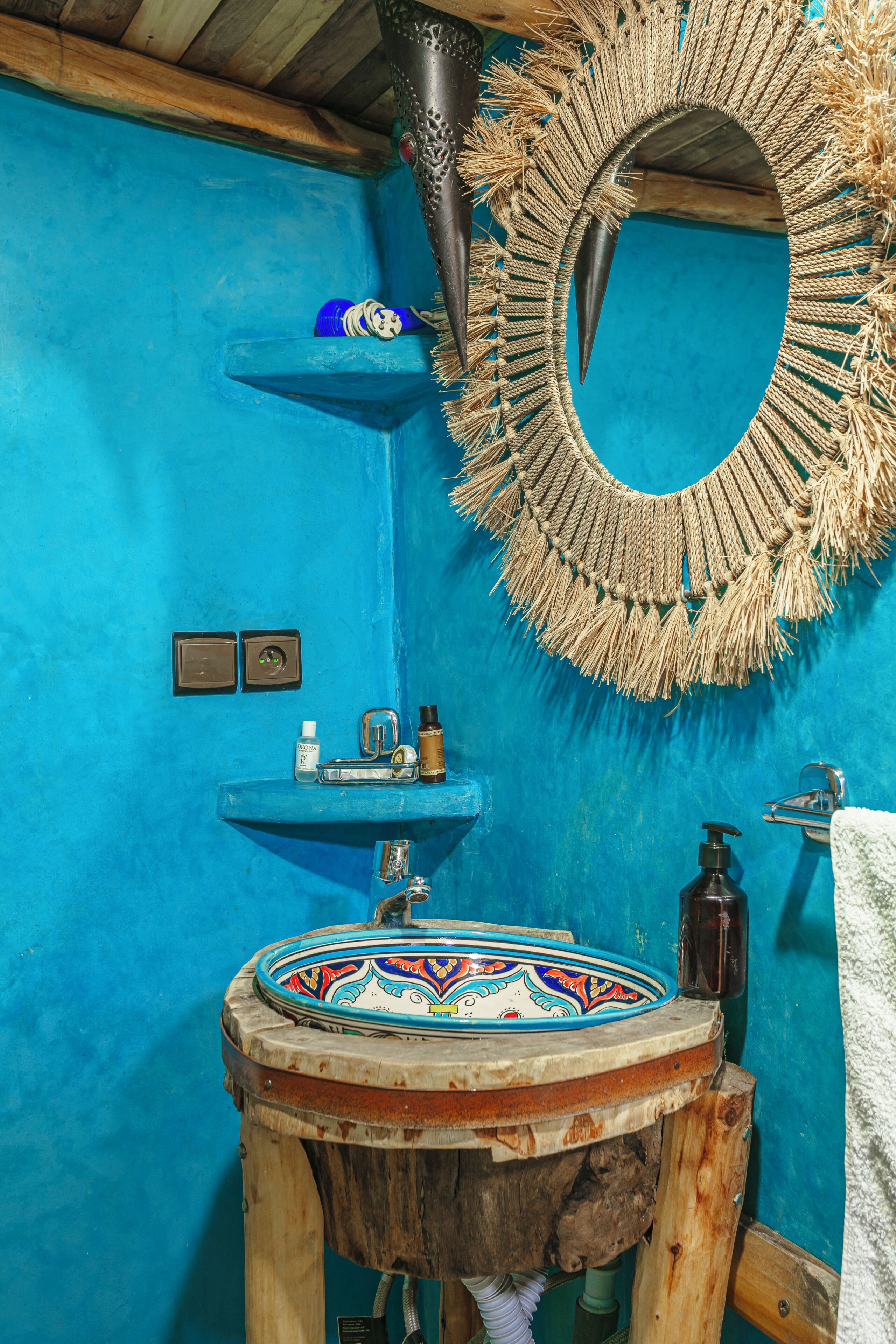 A bathroom with a blue wall, a round mirror with a fringe, a wooden stool with a decorative basin, and a small shelf with toiletries.