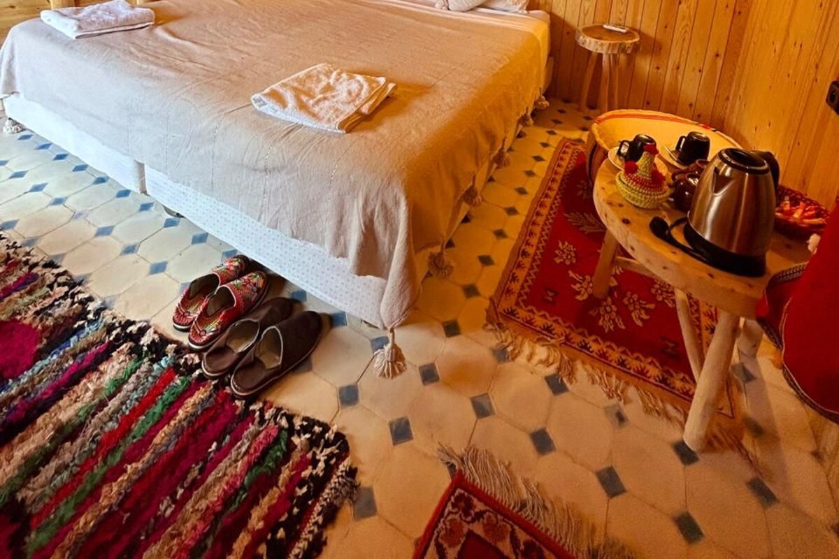 Una habitación con piso de madera, una cama, una mesita con una tetera y una alfombra.