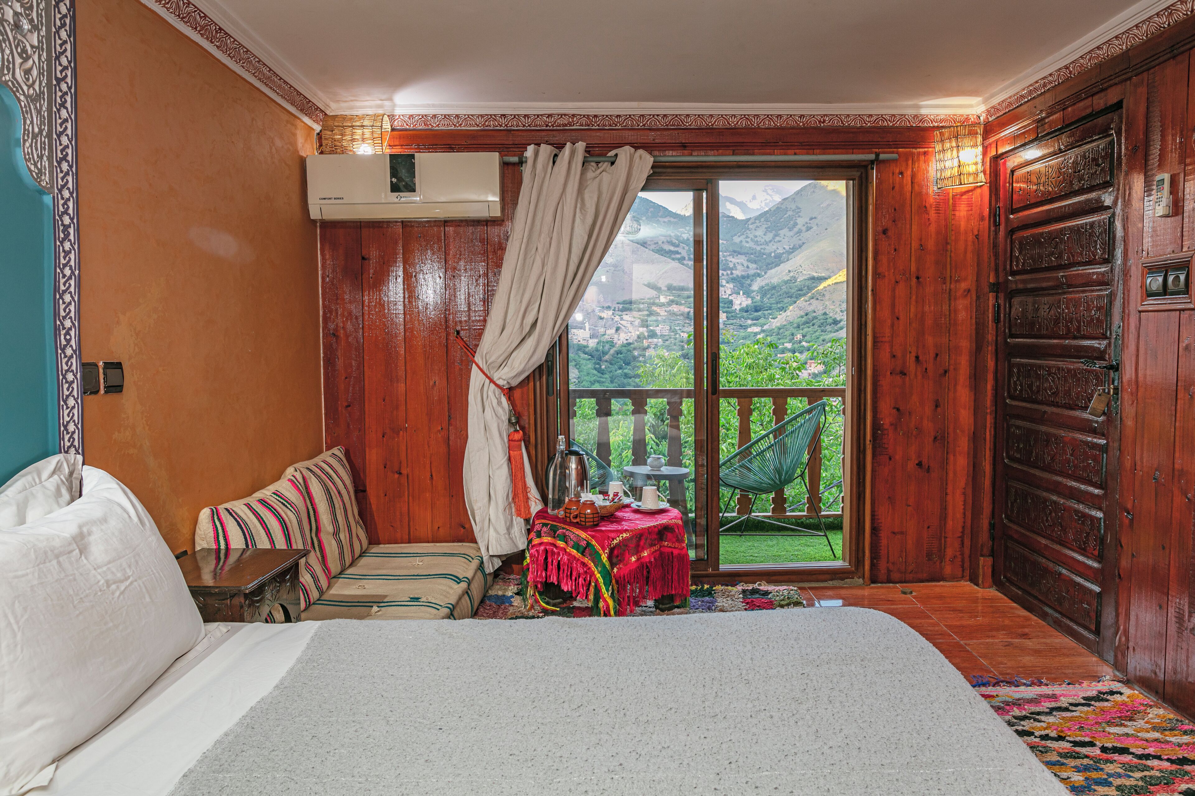A bedroom with a bed, a small table, a chair, and a view of mountains through a large window.
