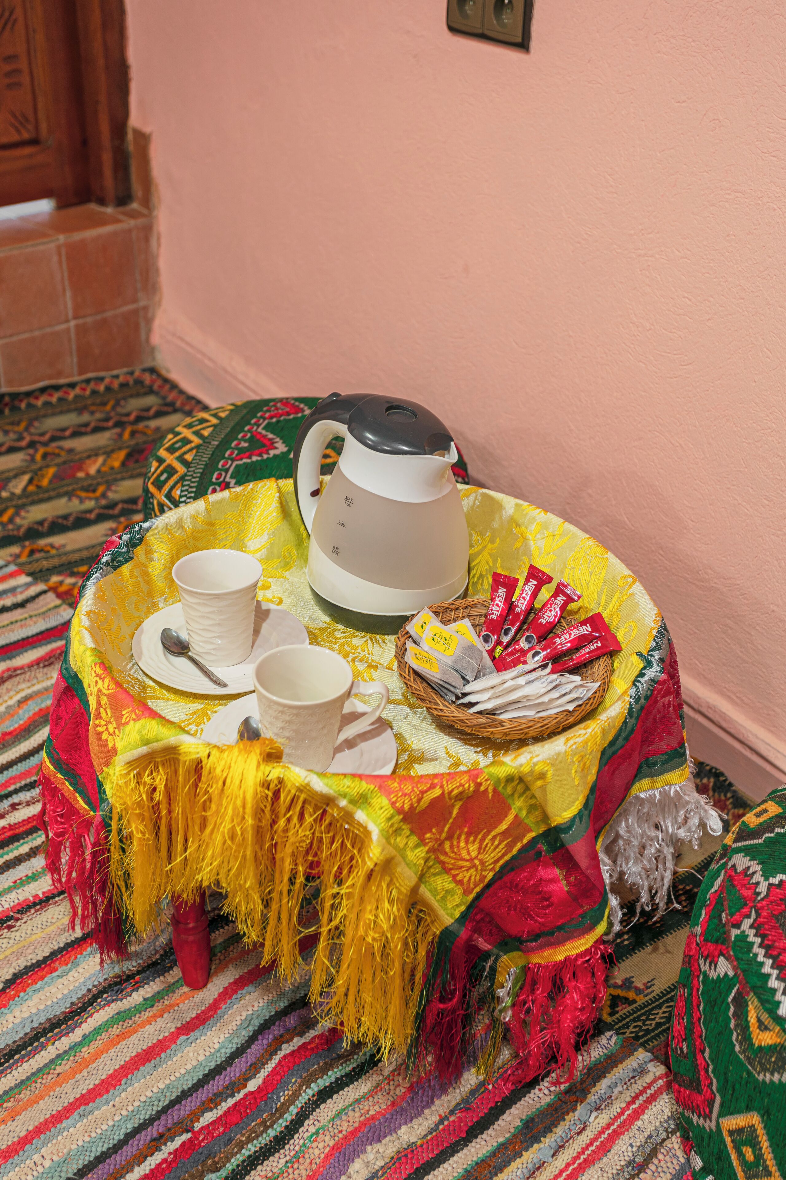 A small round table with a white coffee pot, cups, and a basket of red items, set on a colorful woven rug.