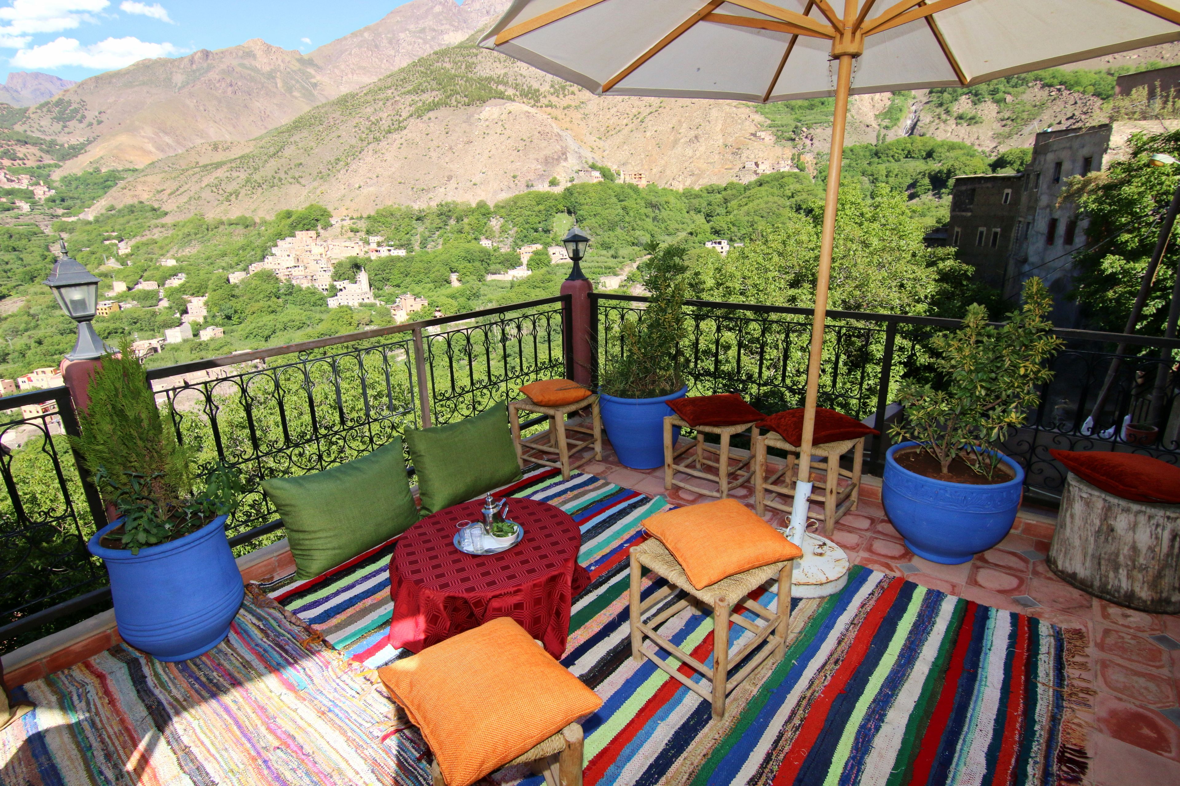 A balcony with a view of a mountainous village, featuring a striped rug, wooden furniture, and potted plants.
