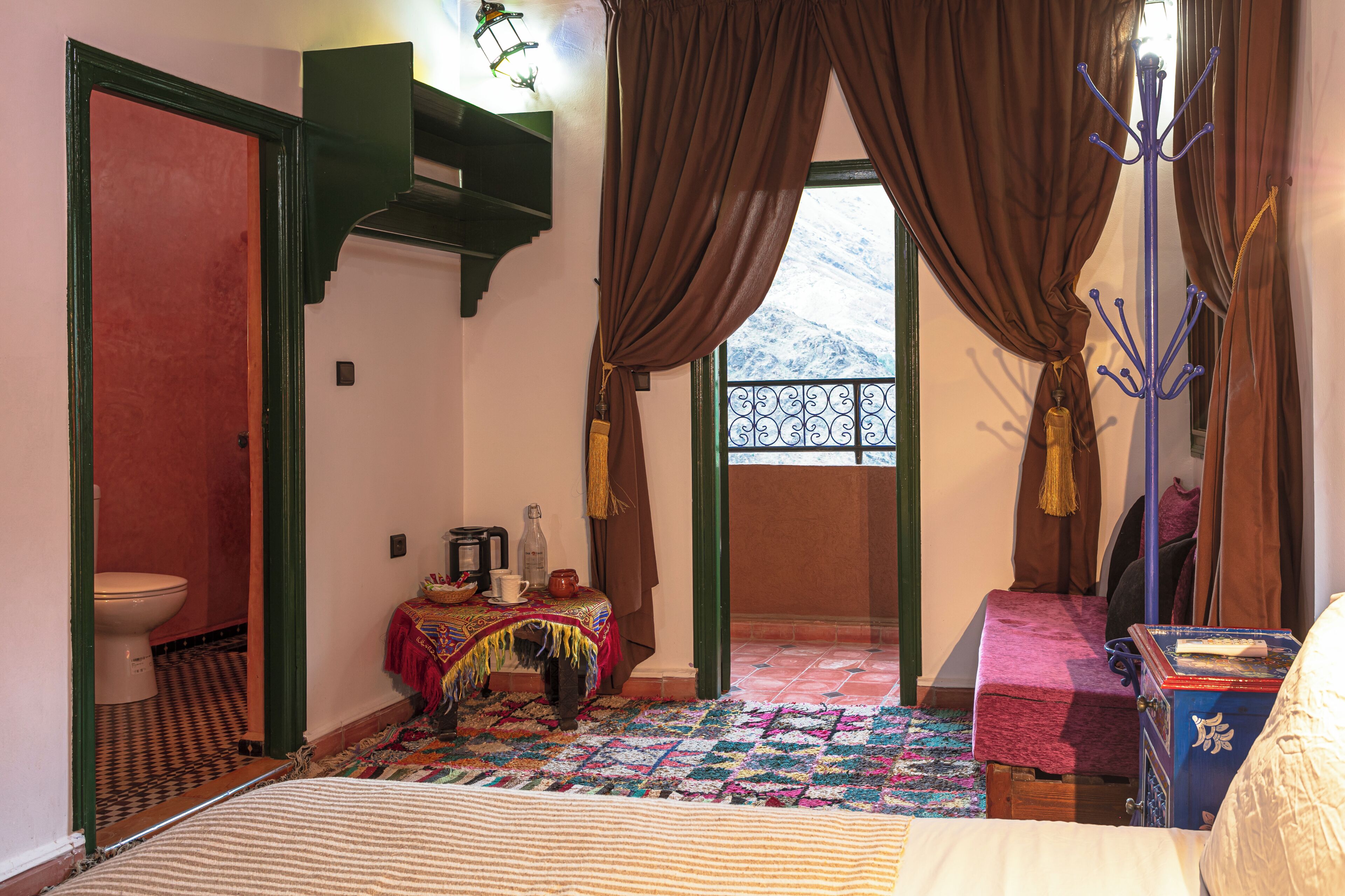 A room with a bed, a table with a basket, a chair, a coat rack, and a view through a window.