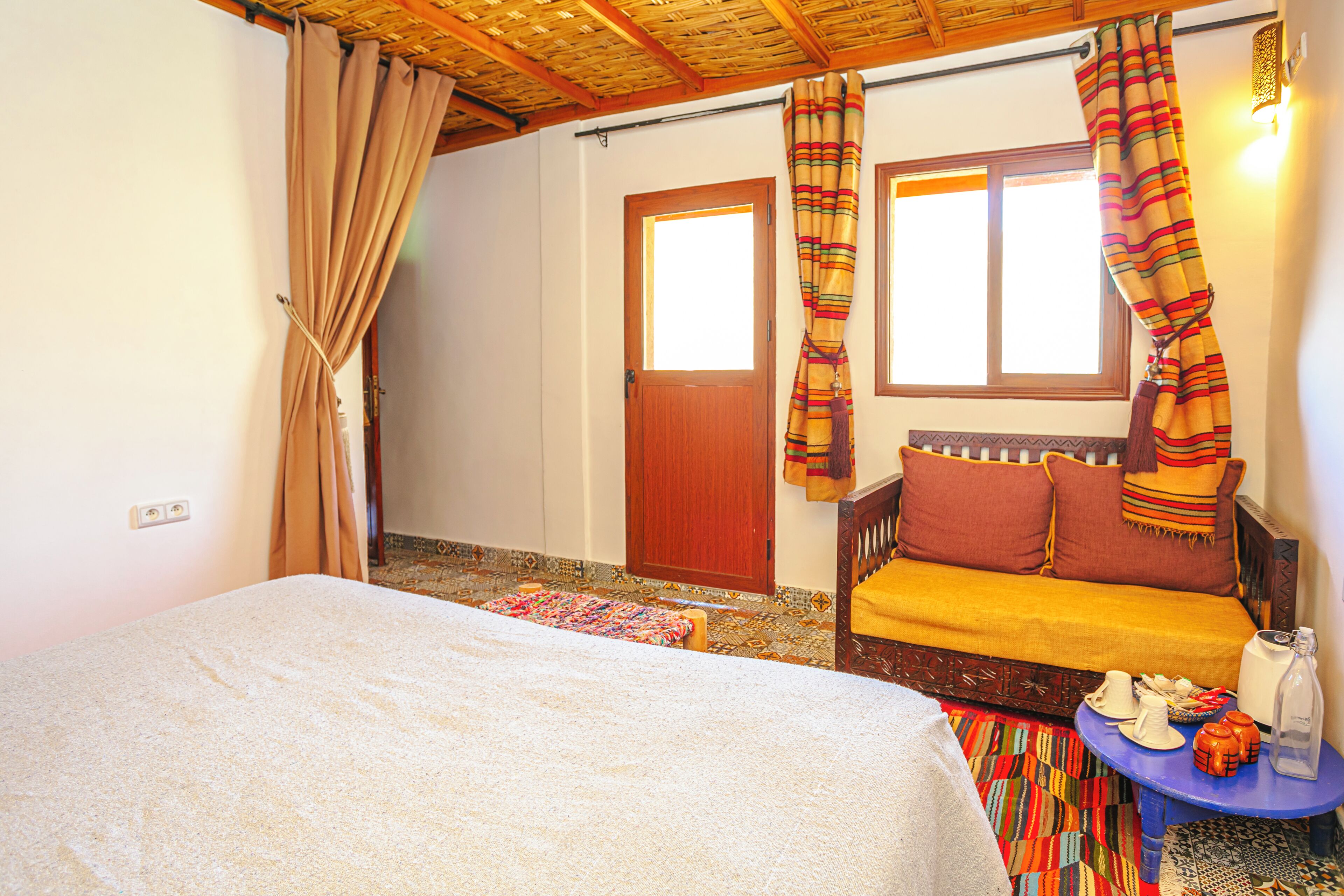 A room with a bed, a sofa, a small table with items, a window, and a door.