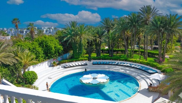 A circular pool with a central fountain, surrounded by lounge chairs and palm trees.