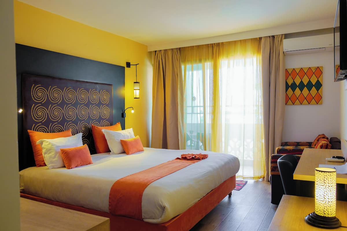 Quarto de hotel com uma cama grande, uma escrivaninha, uma cadeira, um abajur e vista para o exterior.