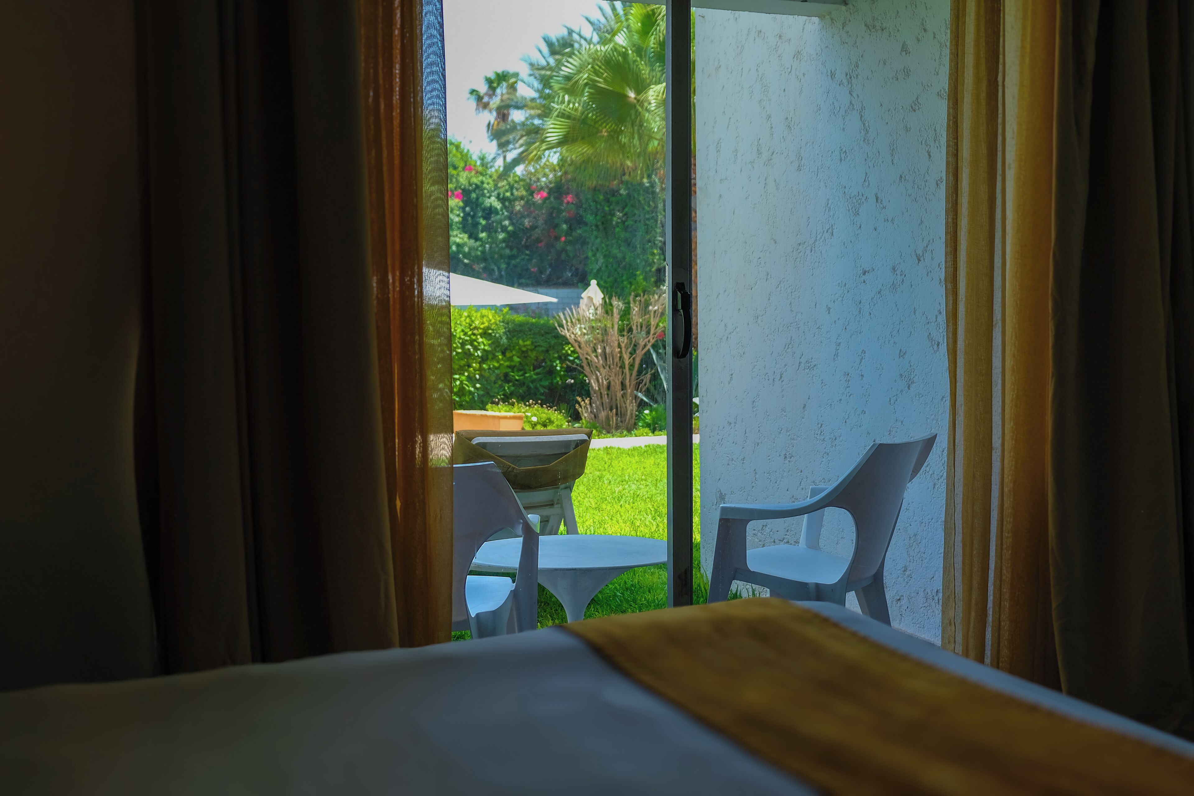 Quarto com cama, janela com cortinas e vista para área externa com mesa e cadeiras.