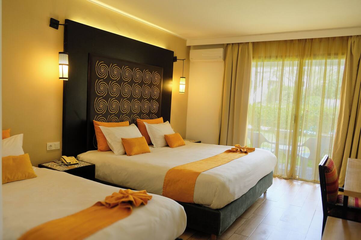 Quarto de hotel com duas camas, cabeceira com painel decorativo, abajures, telefone e vista para o verde pela janela.