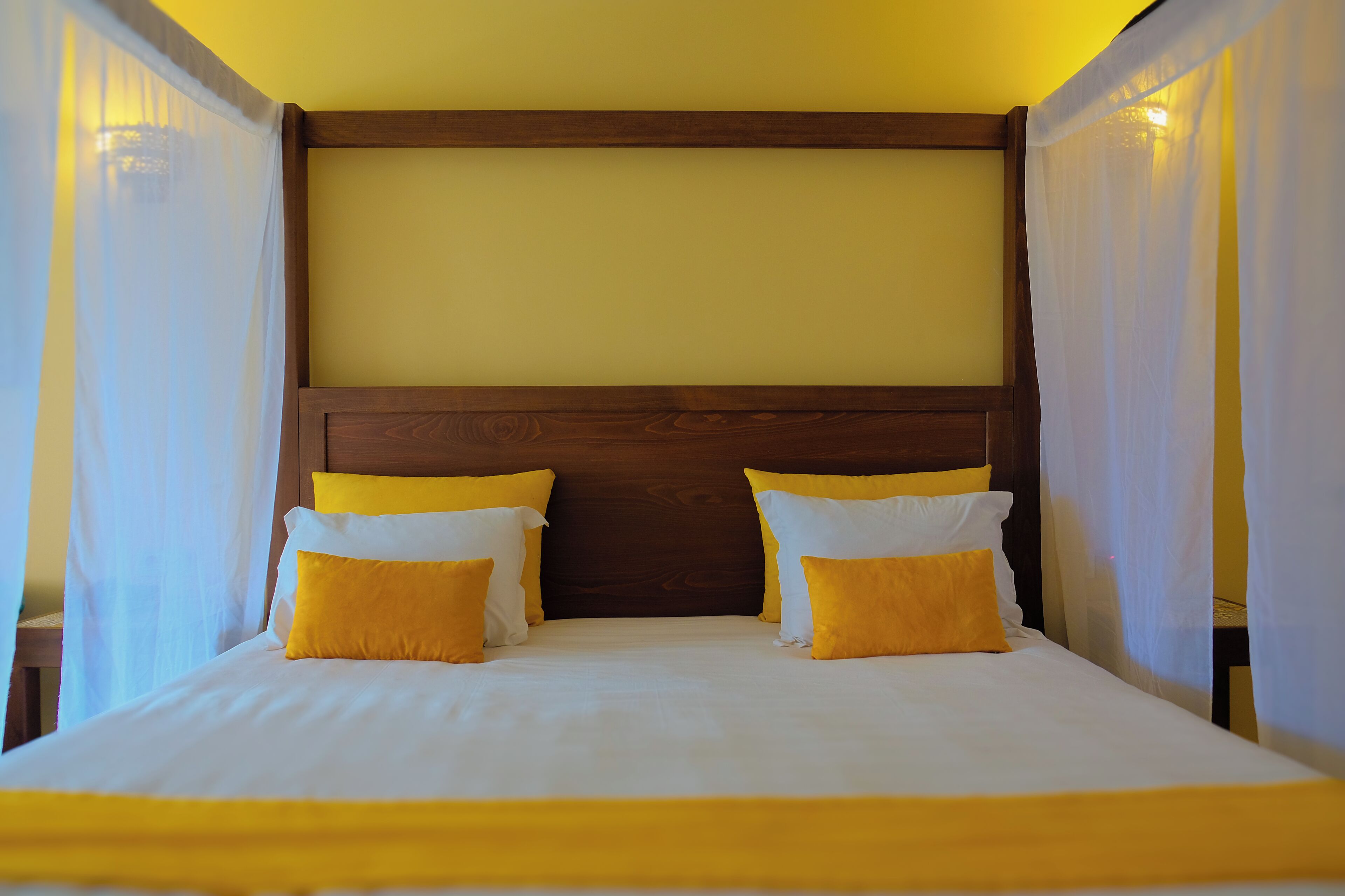 A bed with white and yellow bedding, a wooden headboard, and sheer curtains.