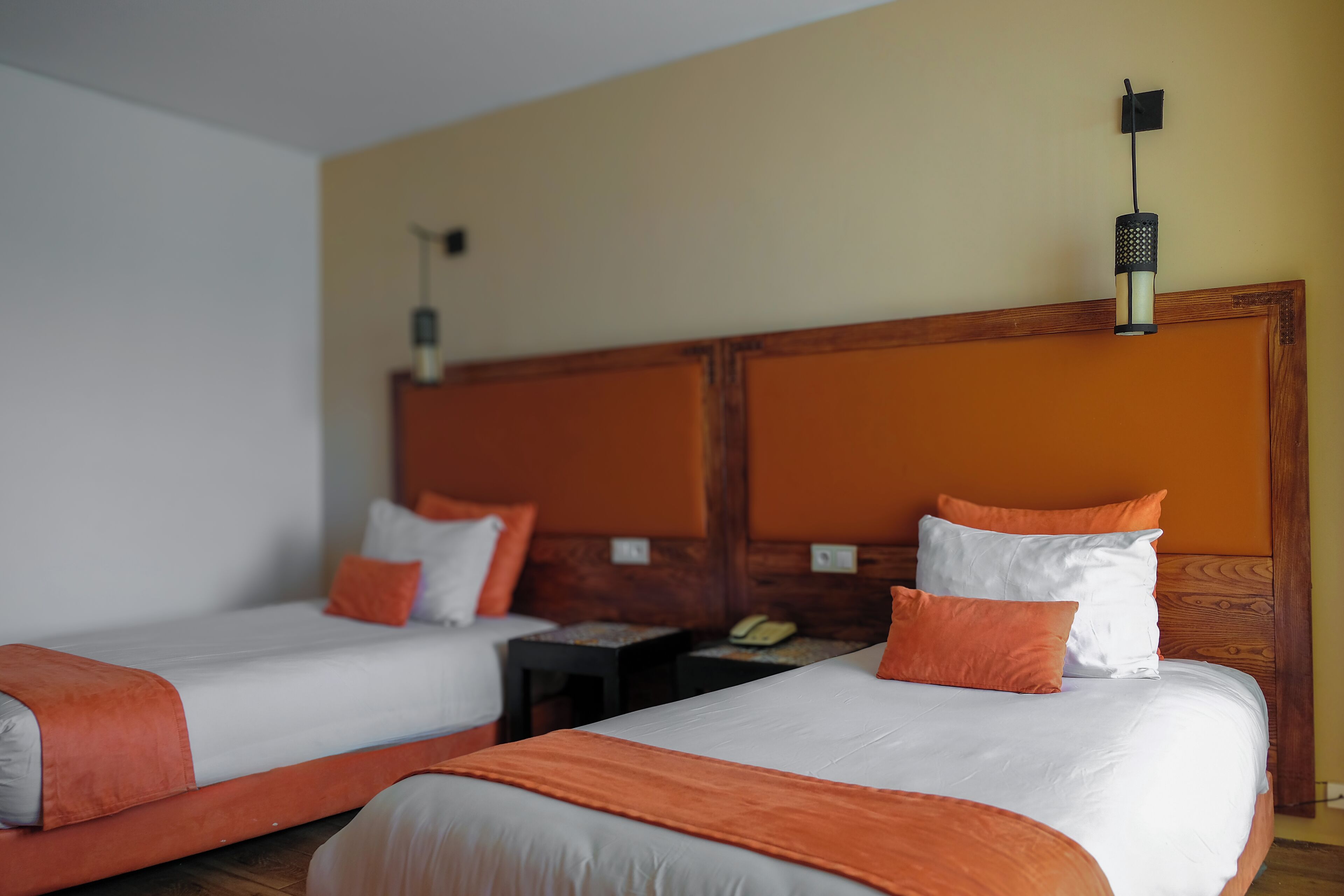 Two single beds with orange bedding, a wooden headboard, a bedside table with a telephone, and a wall-mounted light fixture.