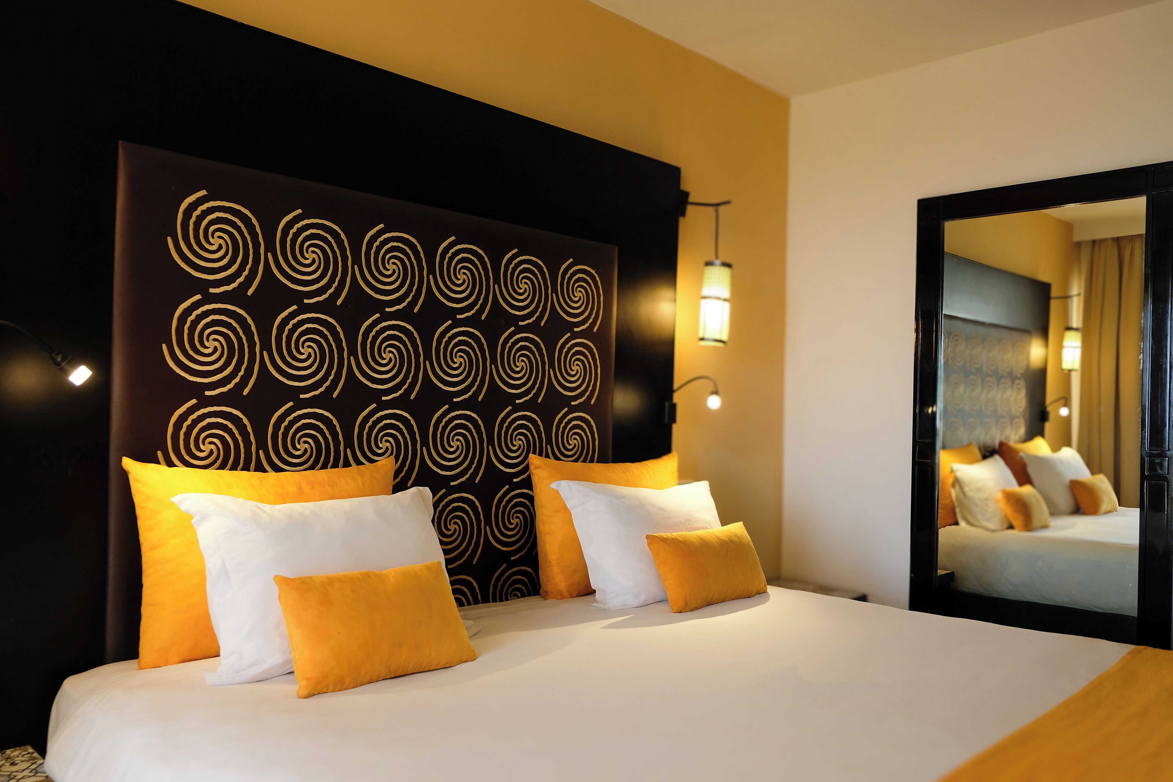Quarto de hotel com uma cama grande, várias almofadas, cabeceira com padrão espiral, luminária de parede e um espelho.