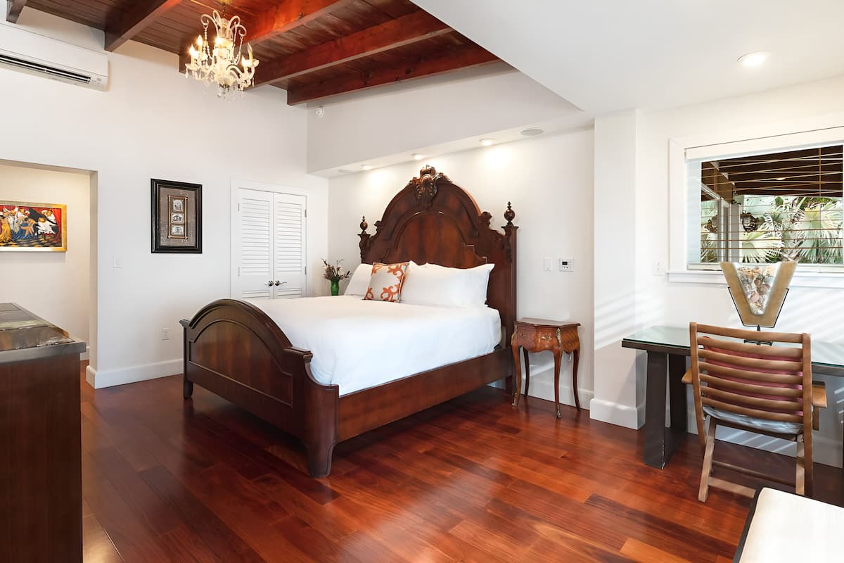 A bedroom with a wooden bed, a chandelier, a desk, and a window with a view of palm trees.