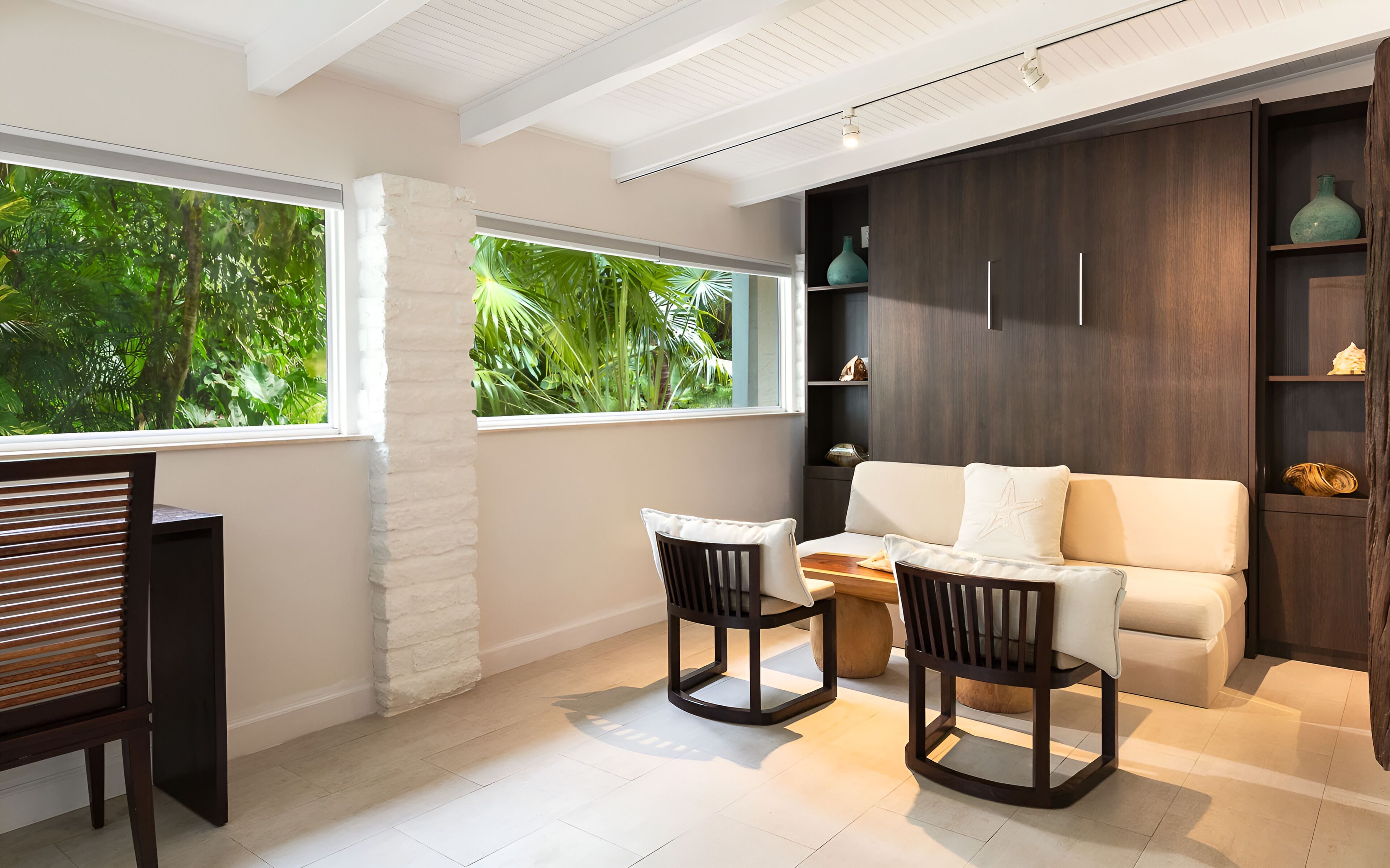 Uma sala de estar moderna com sofá, duas poltronas, mesa de centro e estante de livros. Janelas amplas oferecem vista para o verde lá fora.
