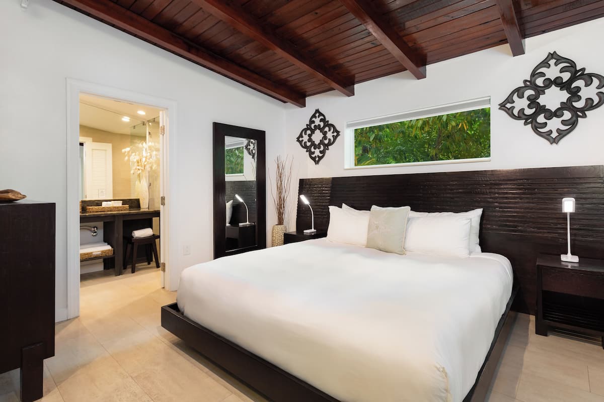 A bedroom with a large bed, a nightstand, a dresser, a mirror, and a window with a view of greenery.