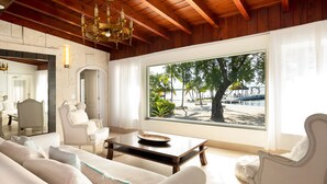 A spacious living room with a large window offering a view of a beach and palm trees. - Largo Resort (Key Largo)