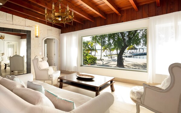 A spacious living room with a large window offering a view of a beach and palm trees. - Largo Resort (Key Largo)