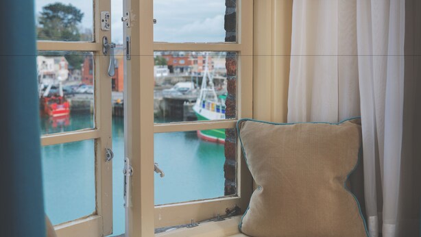 Una habitación con ventana que da a un puerto, un cojín en el alféizar y una cortina entreabierta.