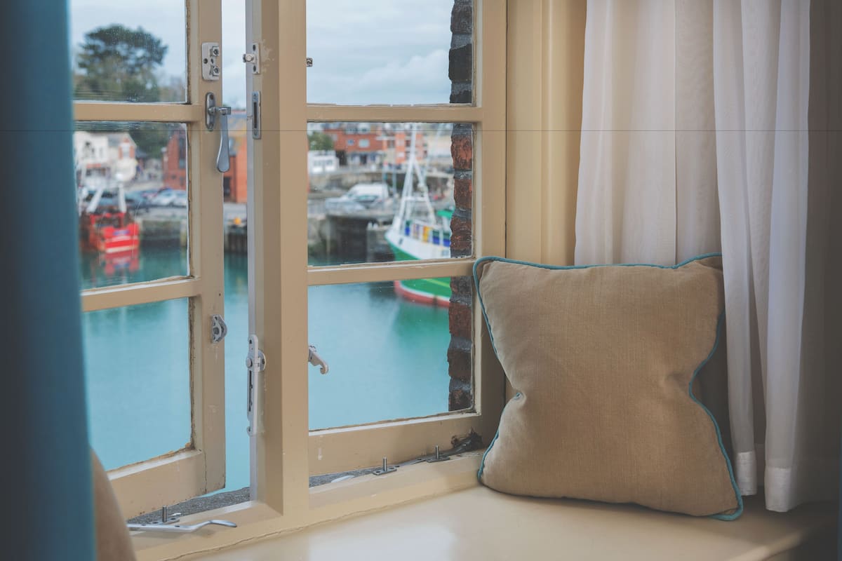 A room with a window overlooking a harbor, a cushion on the windowsill, and a curtain partially drawn.