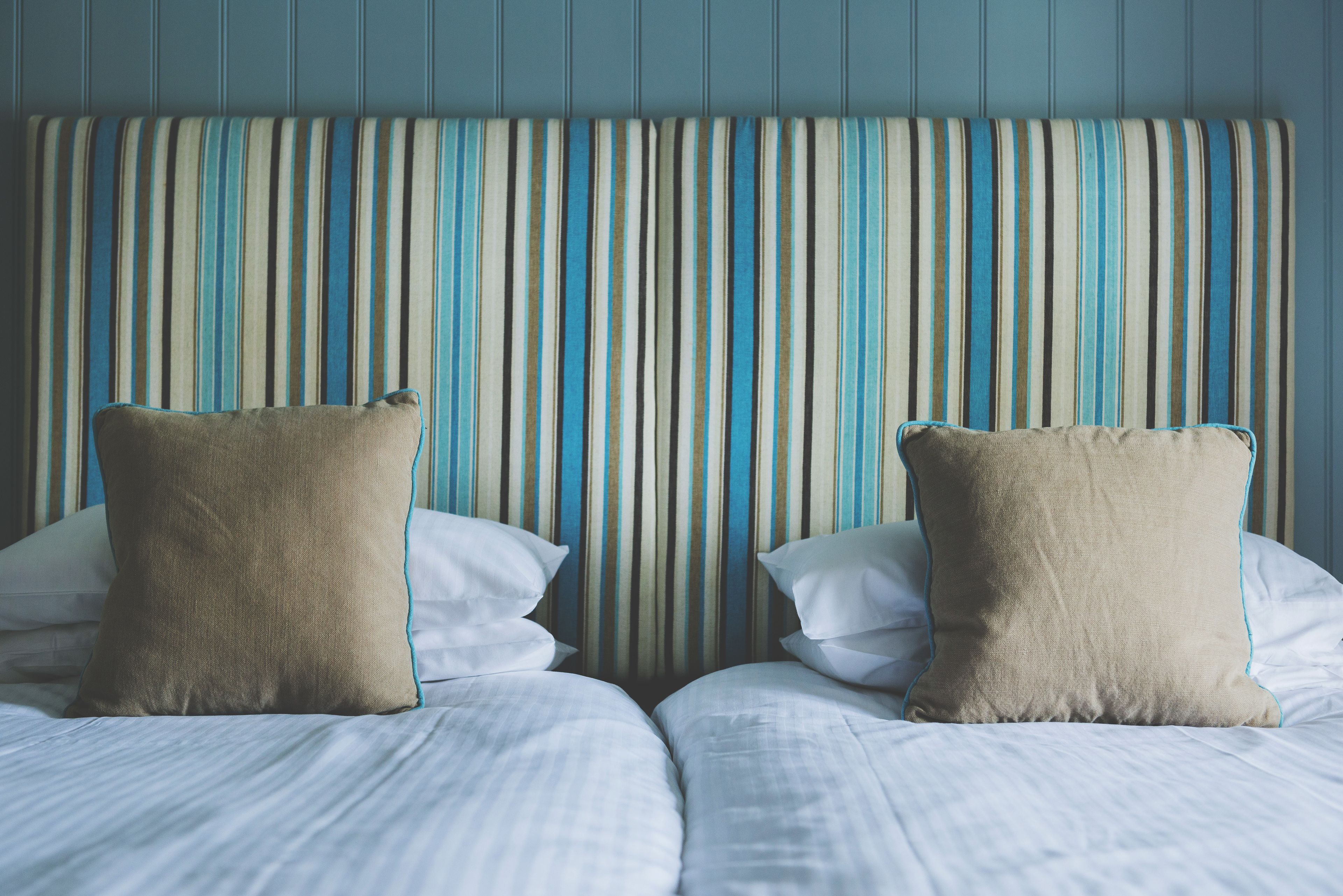 Quarto de hotel com cabeceira listrada, duas camas com roupa de cama branca e almofadas bege.
