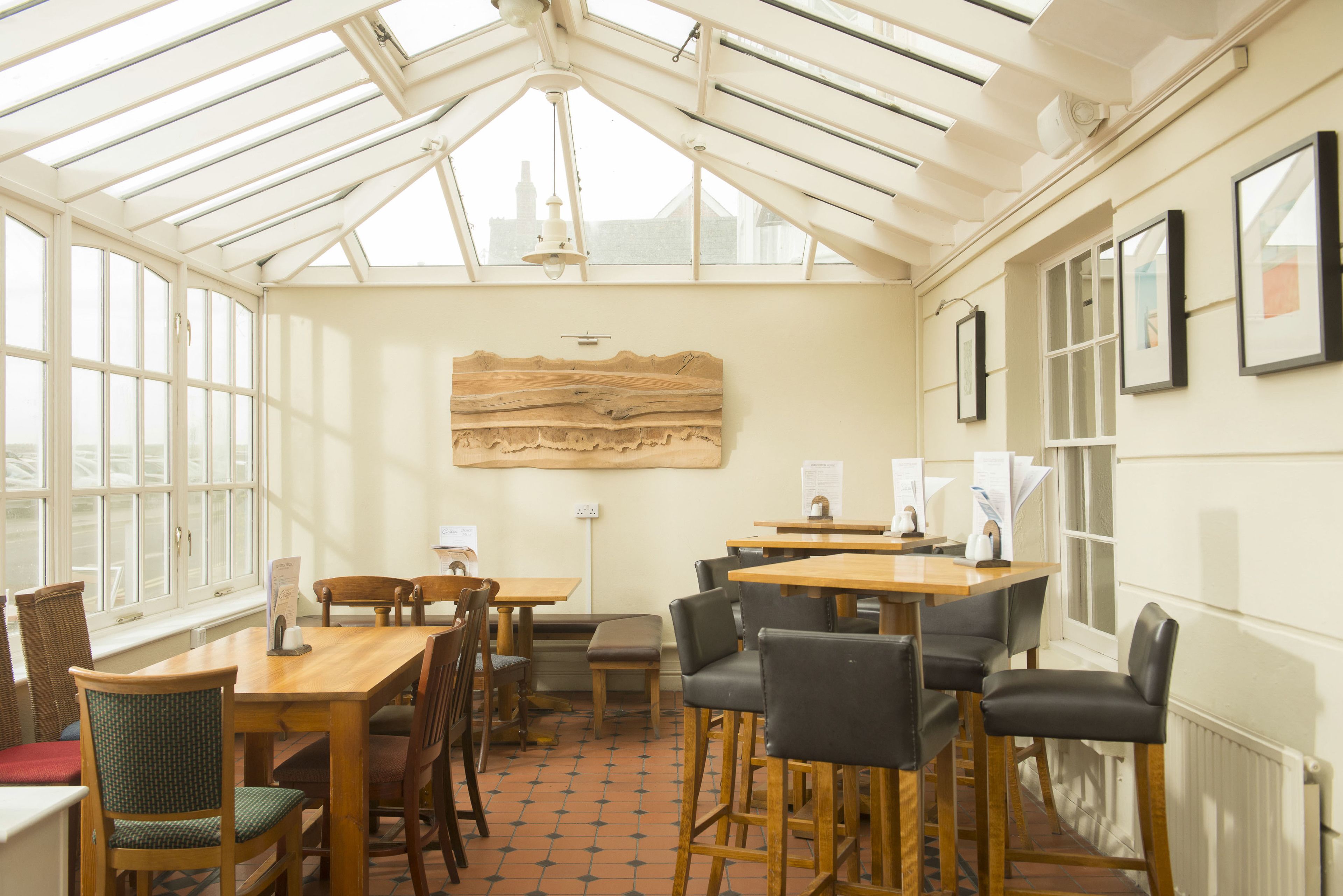 A conservatory with wooden tables and chairs, a large wooden artwork on the wall, and framed pictures on the right.