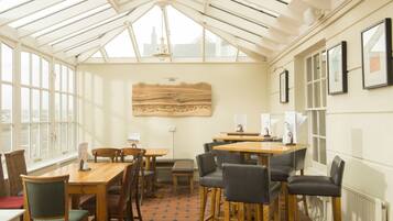 A conservatory with wooden tables and chairs, a large wooden artwork on the wall, and framed pictures on the right.