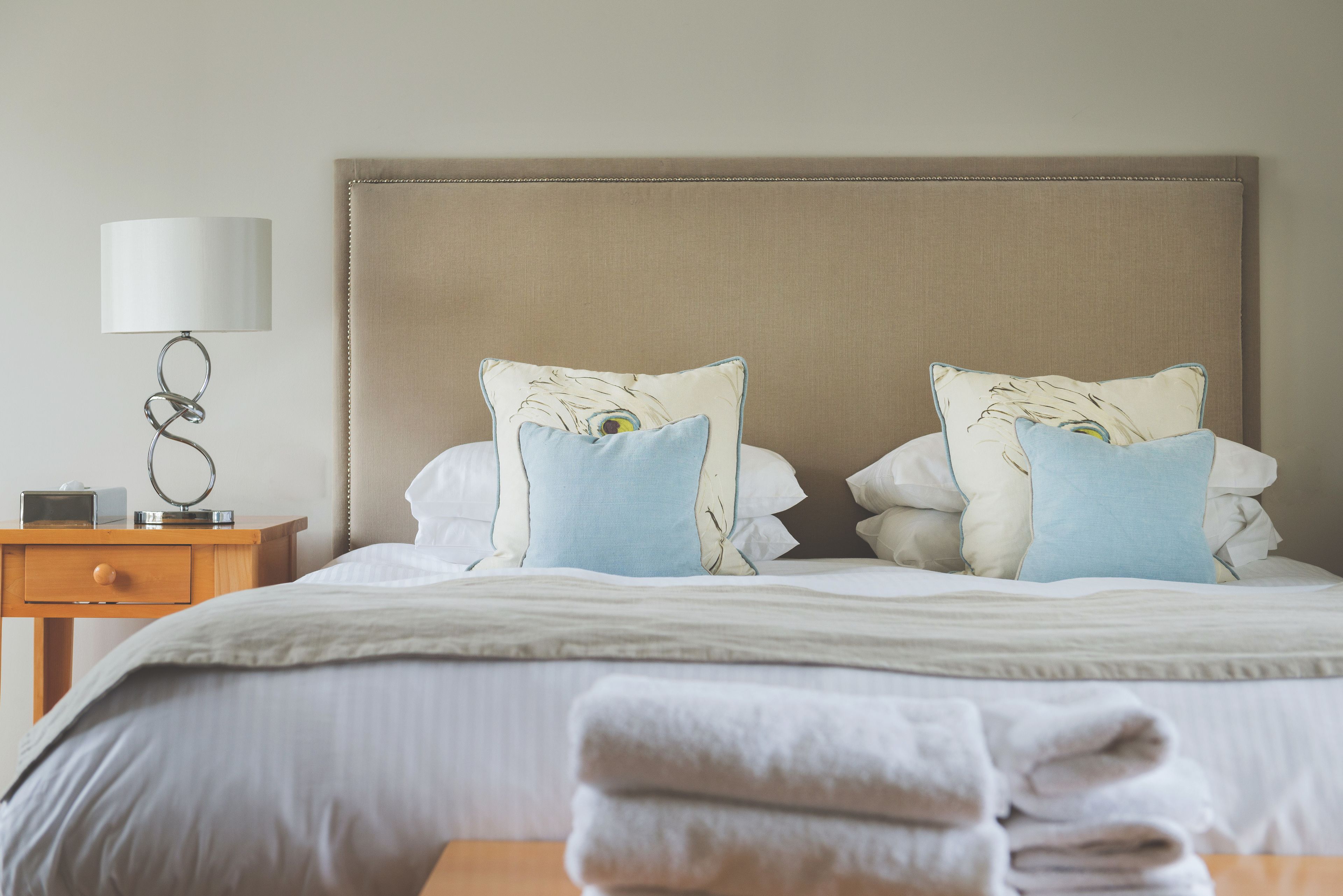 Una cama bien hecha con un cabecero color beige, dos almohadas con estampado de plumas, una lámpara de noche y una mesita de madera con una lámpara y un objeto pequeño encima.
