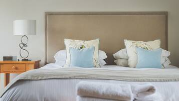 Una cama bien hecha con un cabecero color beige, dos almohadas con estampado de plumas, una lámpara de noche y una mesita de madera con una lámpara y un objeto pequeño encima.