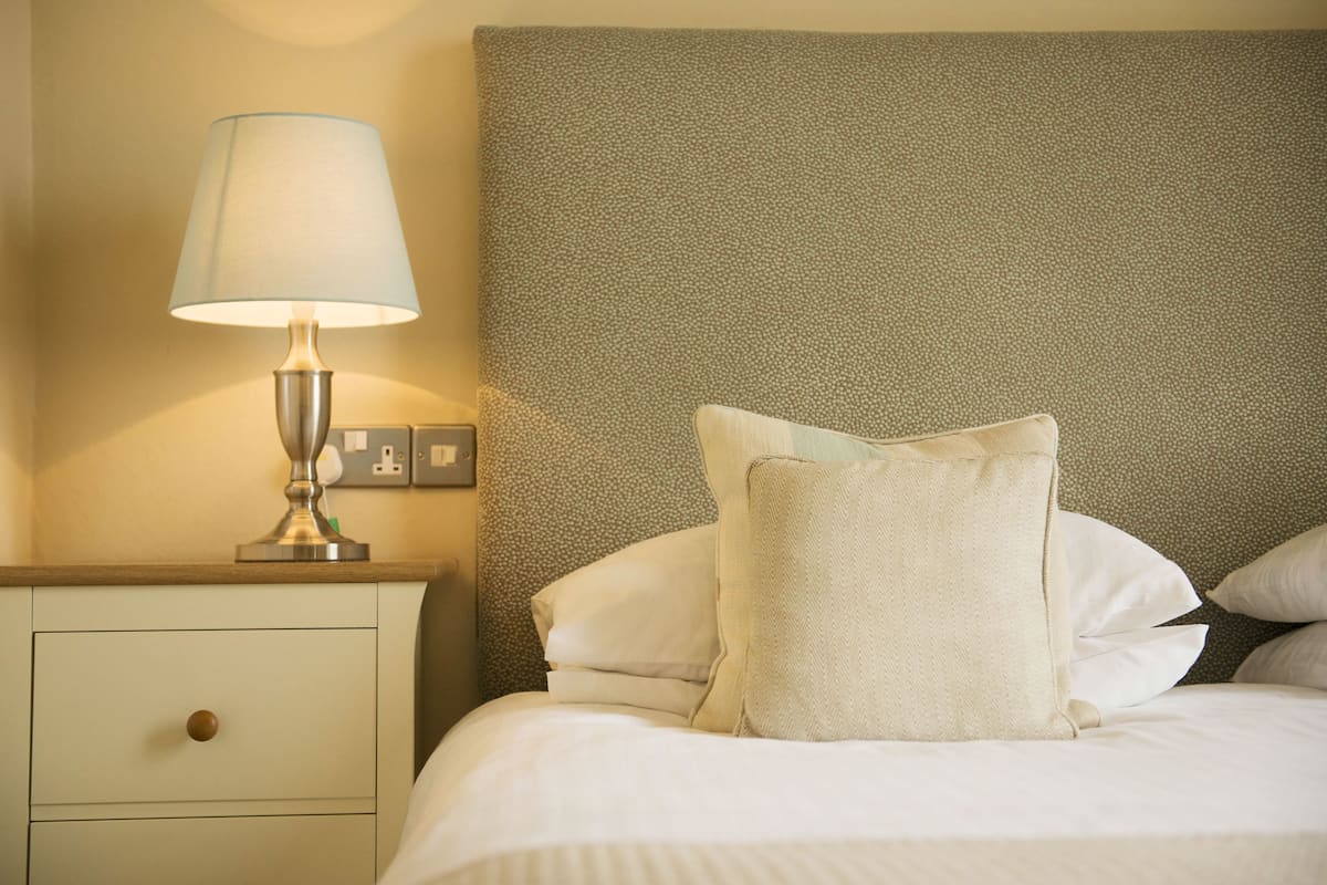 A neatly made bed with white linens and a textured headboard, a bedside lamp, and a small bedside table with a drawer.