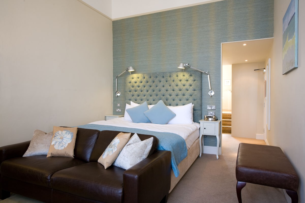 A bedroom with a large bed, a brown leather sofa, a small bench, and a nightstand with a lamp.