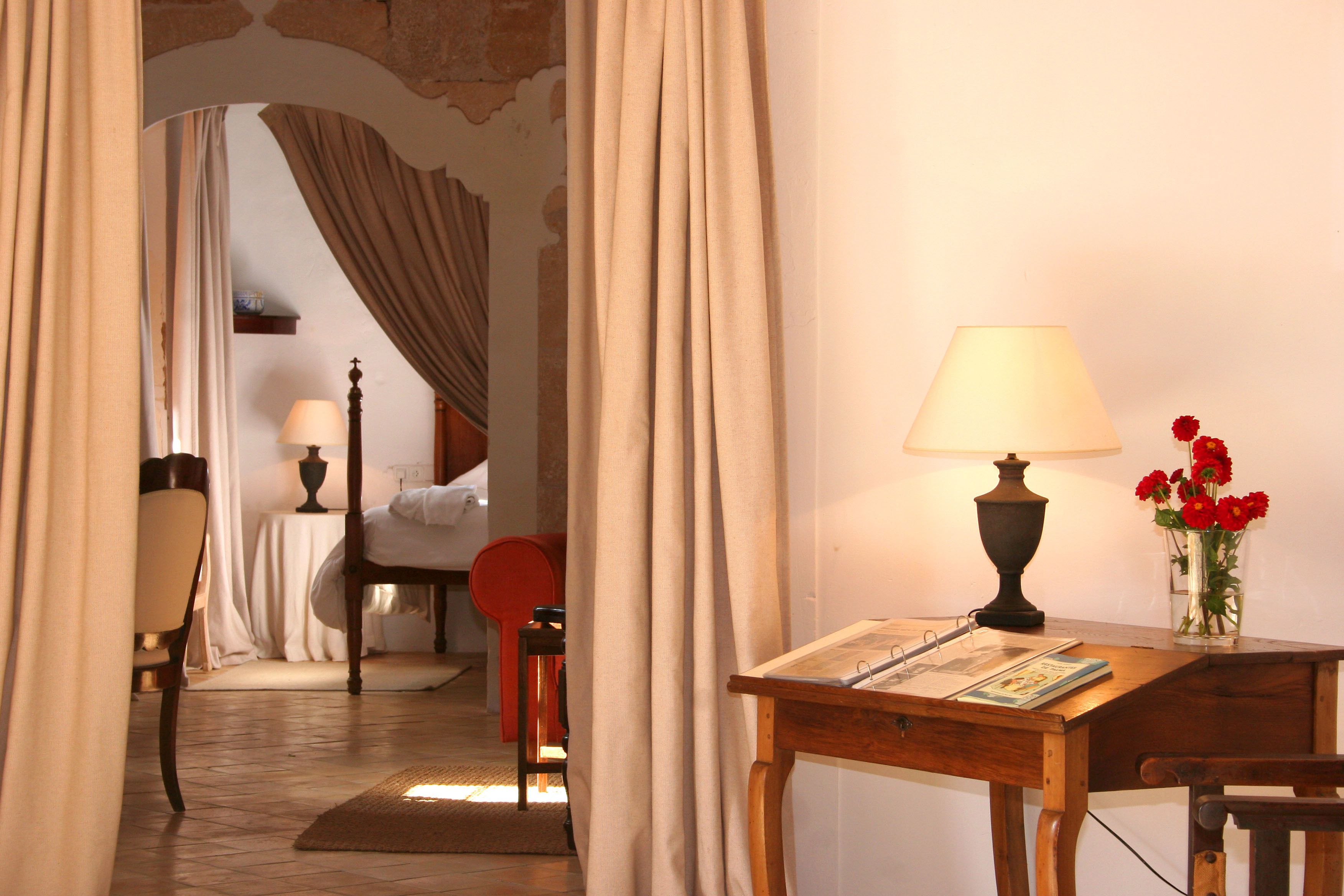A room with a wooden table, a lamp, a vase with flowers, and a bed.