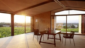 Un comedor espacioso con una mesa dispuesta para cuatro, ventanas amplias que ofrecen vistas a la vegetación y edificios lejanos, y un techo alto con vigas a la vista.