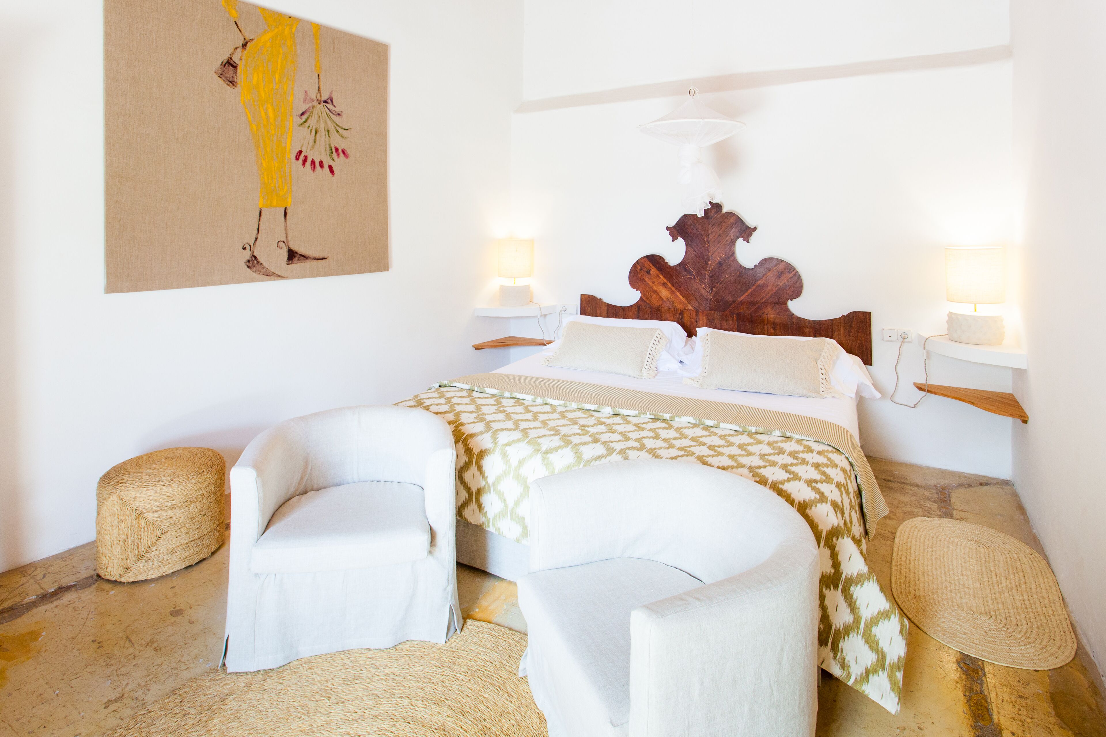 A bedroom with a bed, two bedside tables, two chairs, a wooden headboard, and a wall-mounted artwork.