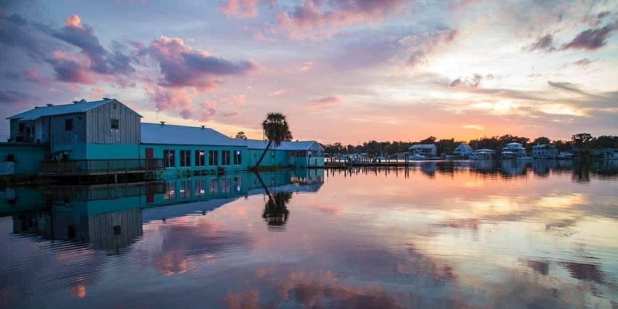 A waterfront building with a reflective water surface, a dock, and a row of houses in the background.