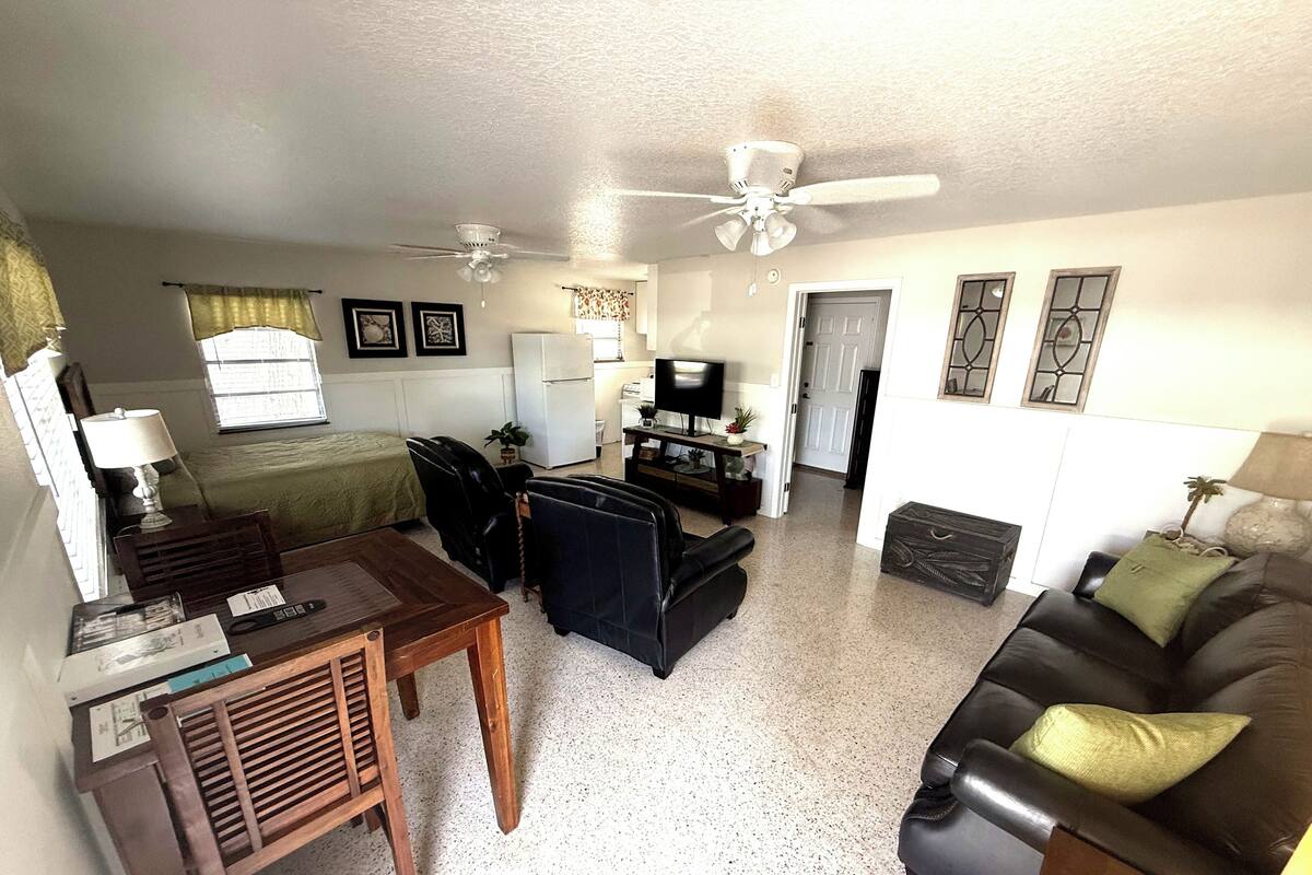 A compact living space with a bed, a dining table, a sofa, and a TV. There are ceiling fans, a window with curtains, and a door leading to another room.